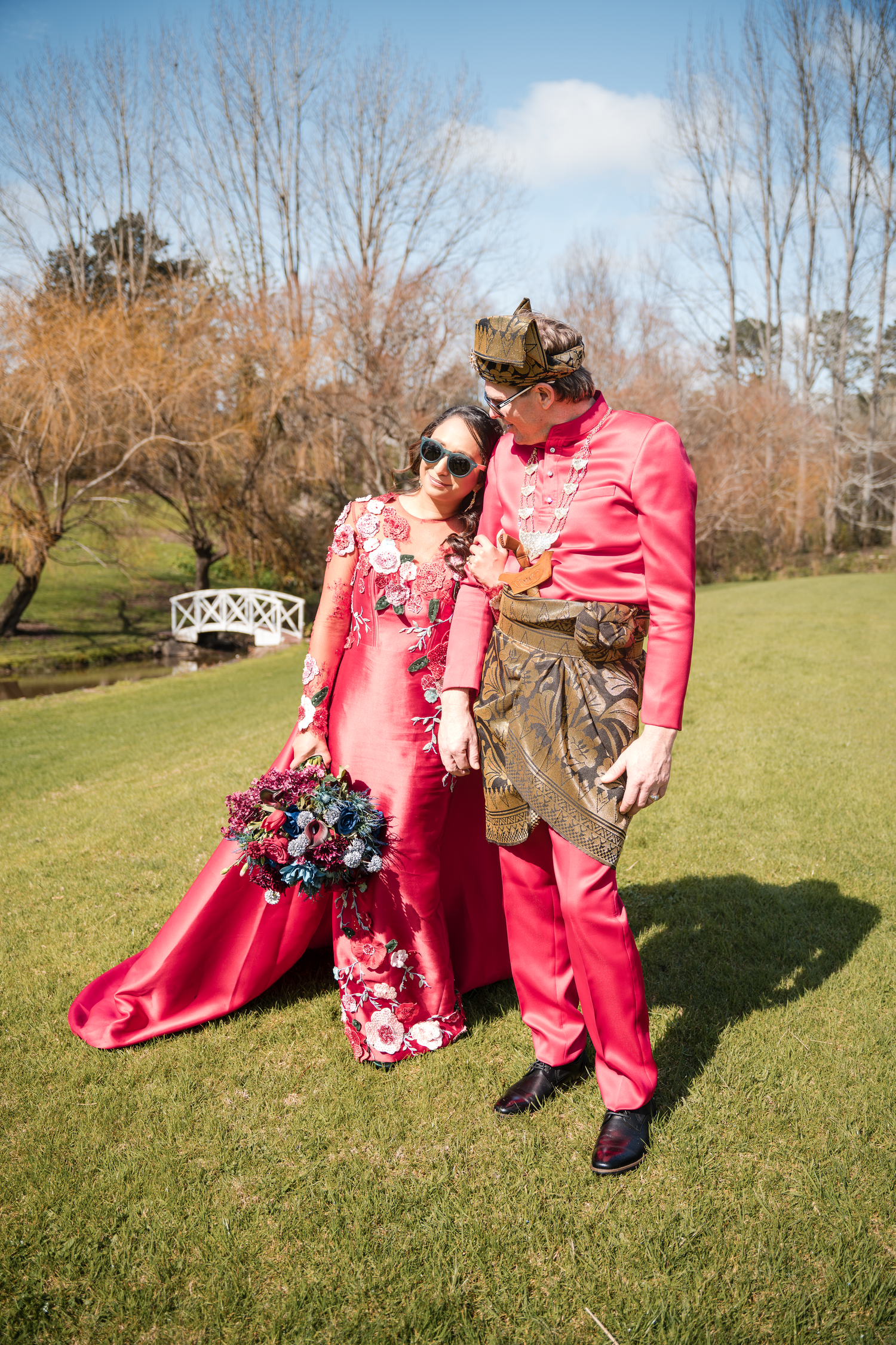 Couple in coordinating pink outfits pose together on manicured lawn with gazebo in background.
