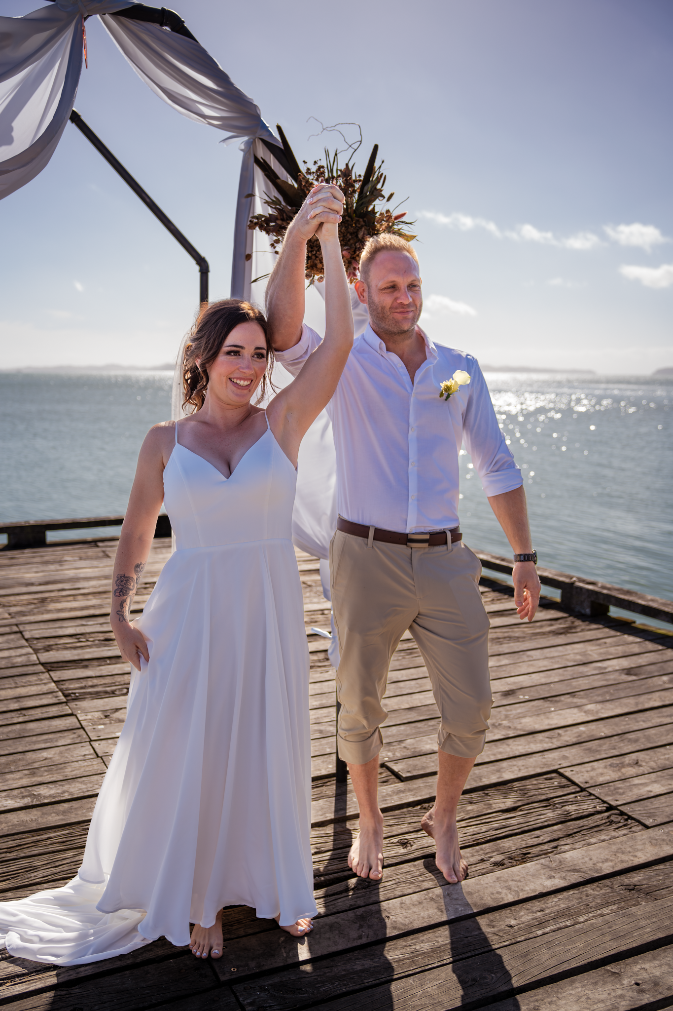 Newlyweds celebrate under a white fabric canopy on a wooden pier by the ocean on their wedding day.