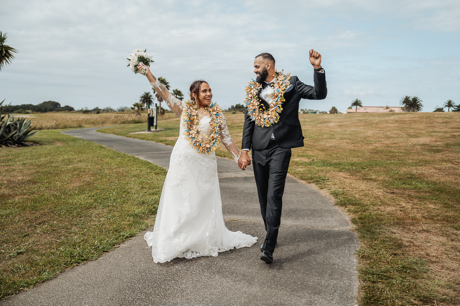 Joyful newlyweds celebrate while walking on pathway with tropical scenery.