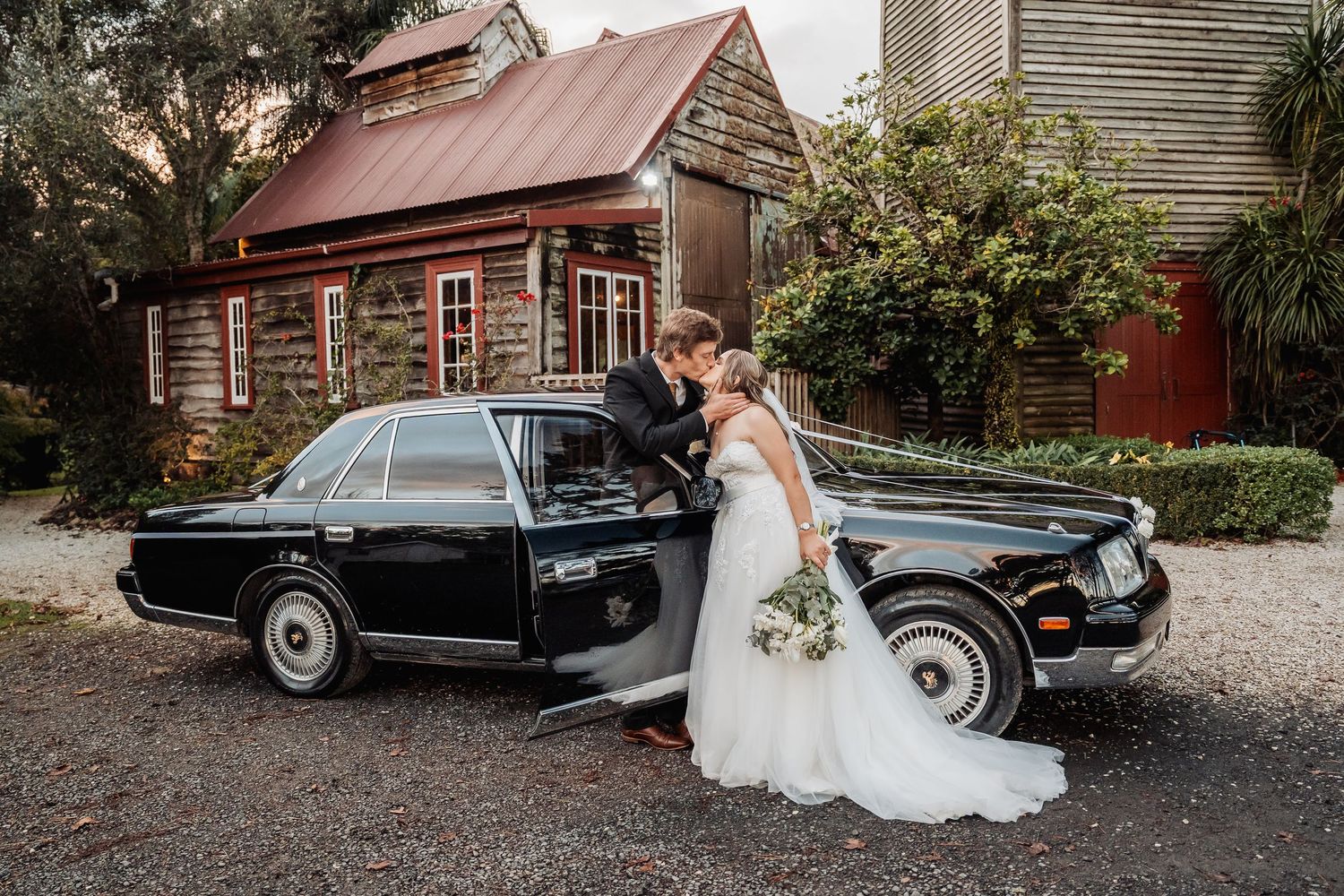 A classic black vintage car parked in front of rustic wooden buildings serves as an elegant wedding backdrop.