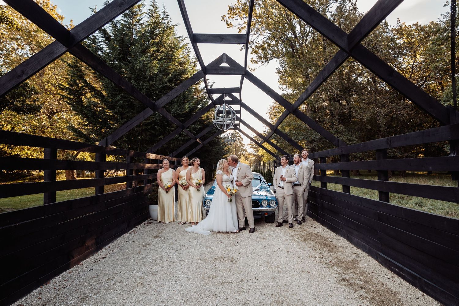 Wedding party posing on black bridge with vintage blue car and autumn trees.