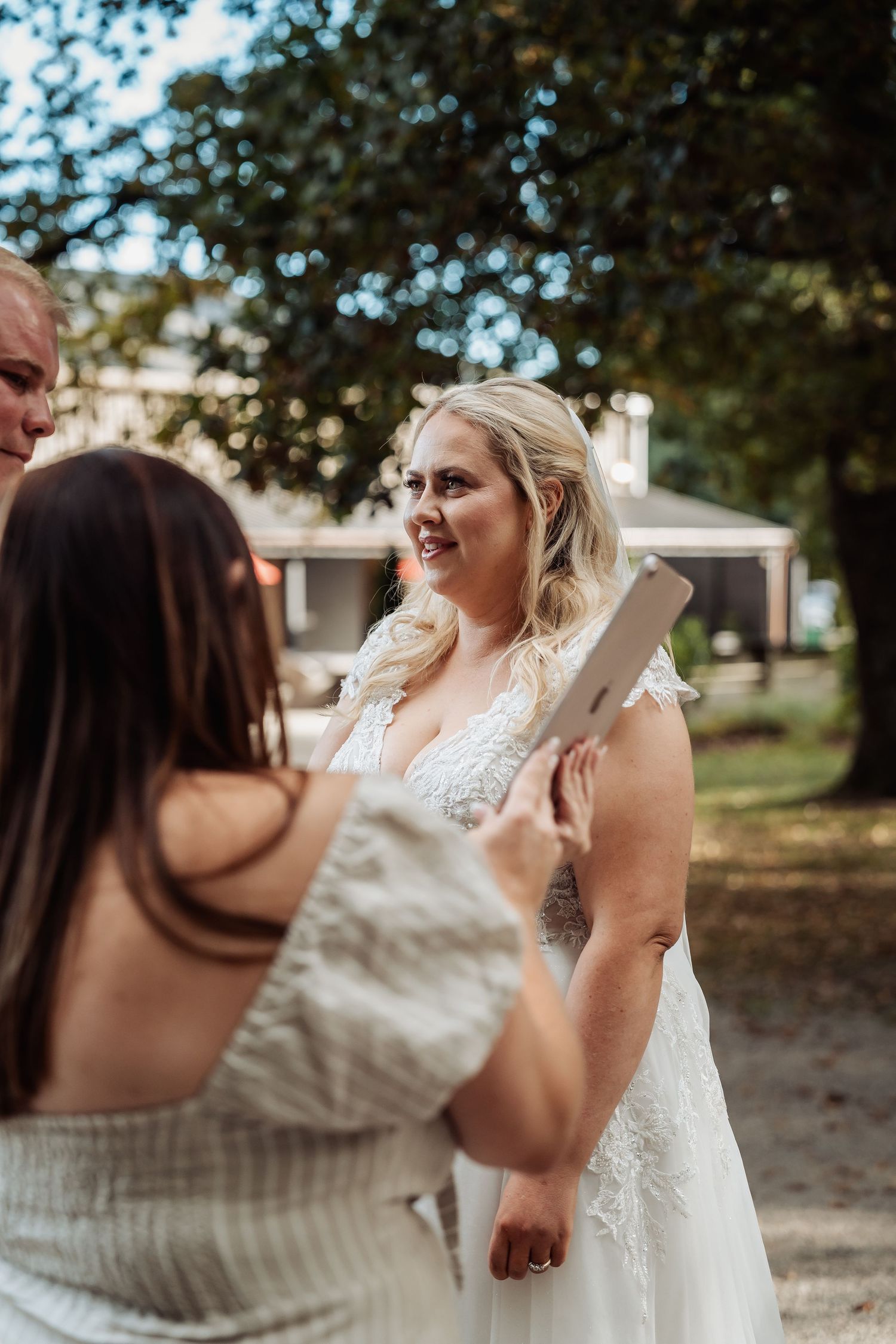 Bride in lace wedding gown smiling during outdoor wedding ceremony exchange with officiant.