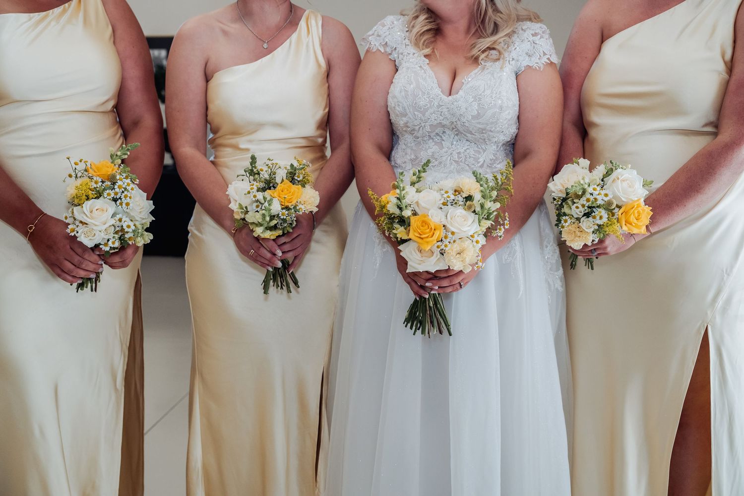 Bride in lace wedding gown poses with three bridesmaids in champagne dresses holding yellow and white bouquets.