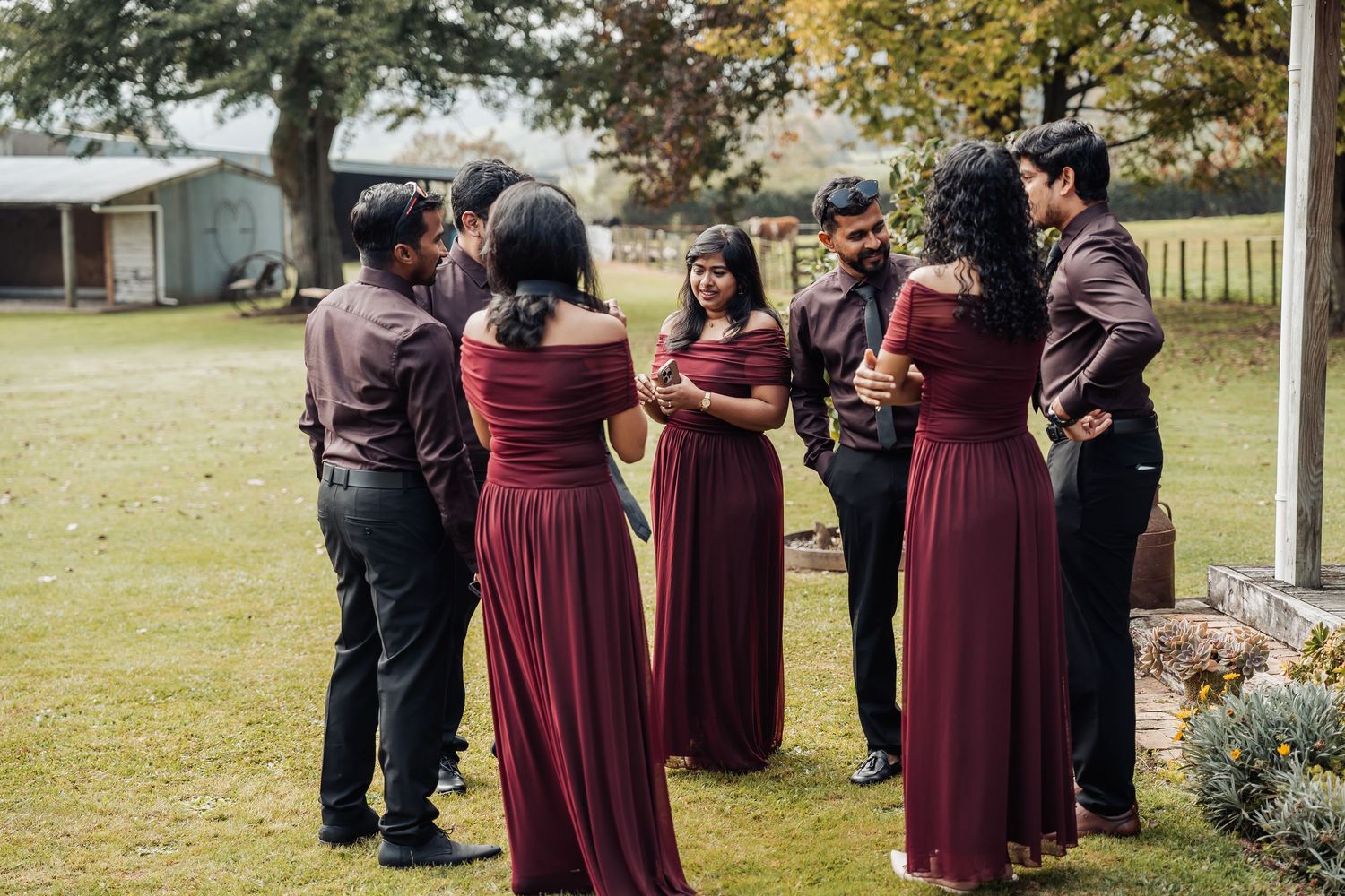 Wedding party in matching burgundy outfits gathered on green lawn, bridesmaids in off-shoulder gowns with groomsmen.