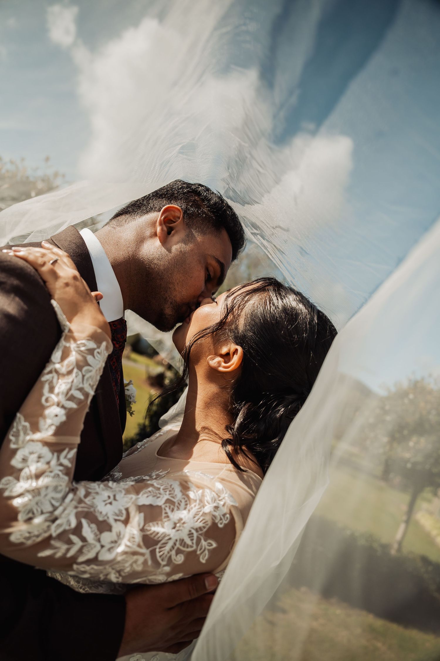 Bride and groom sharing a romantic kiss under flowing veil against a blue sky backdrop.