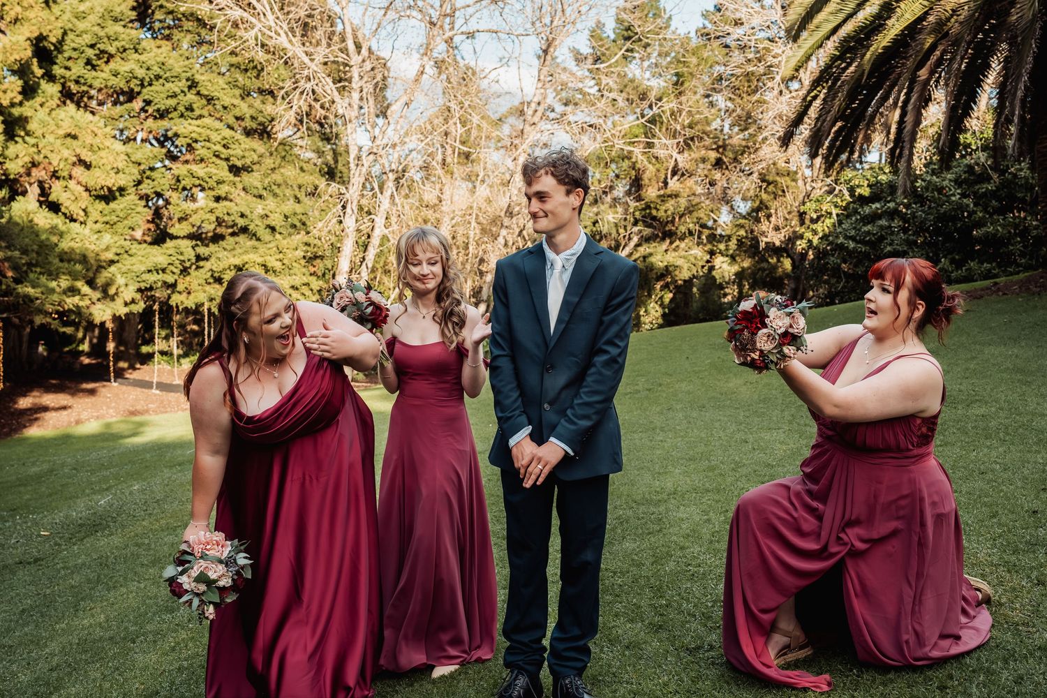 Bridesmaids in burgundy gowns laugh with groomsman on green lawn at wedding.