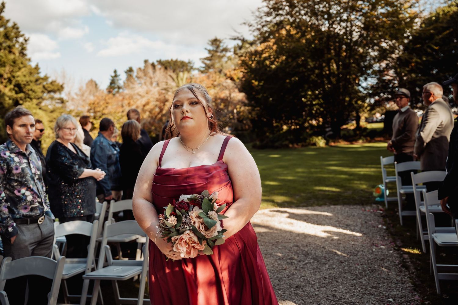 Bridesmaid in burgundy gown walks down outdoor wedding aisle holding floral bouquet with guests seated on white chairs.