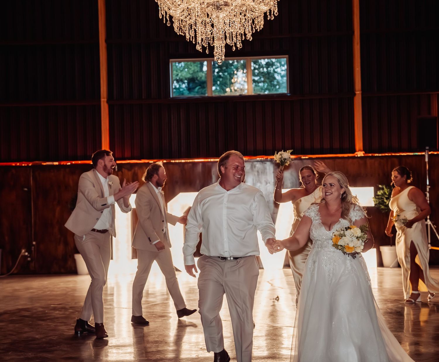 Bride and groom celebrating with wedding party on dance floor under chandelier in rustic barn reception.