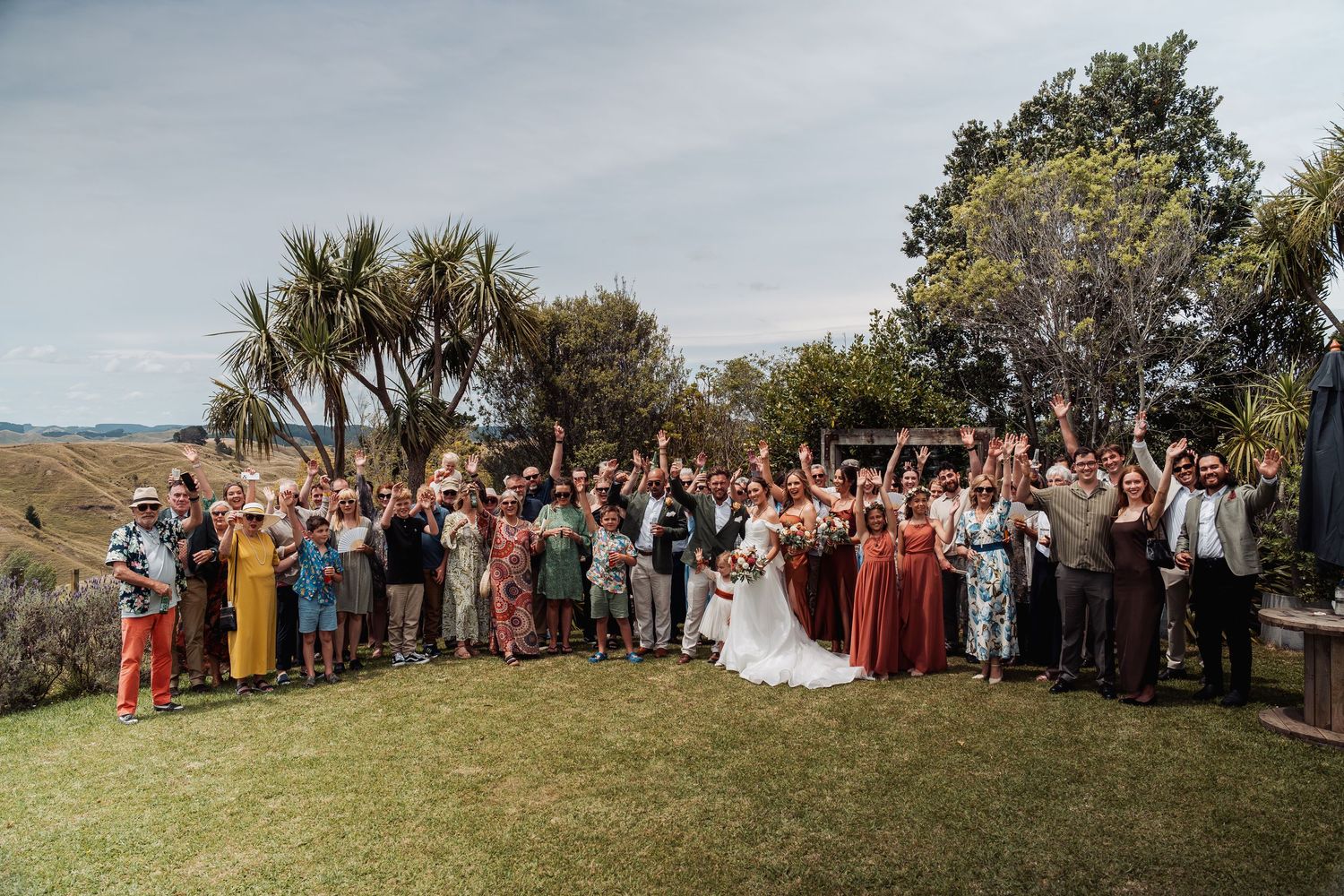 Large wedding group photo with bride and groom surrounded by guests cheering in a garden.