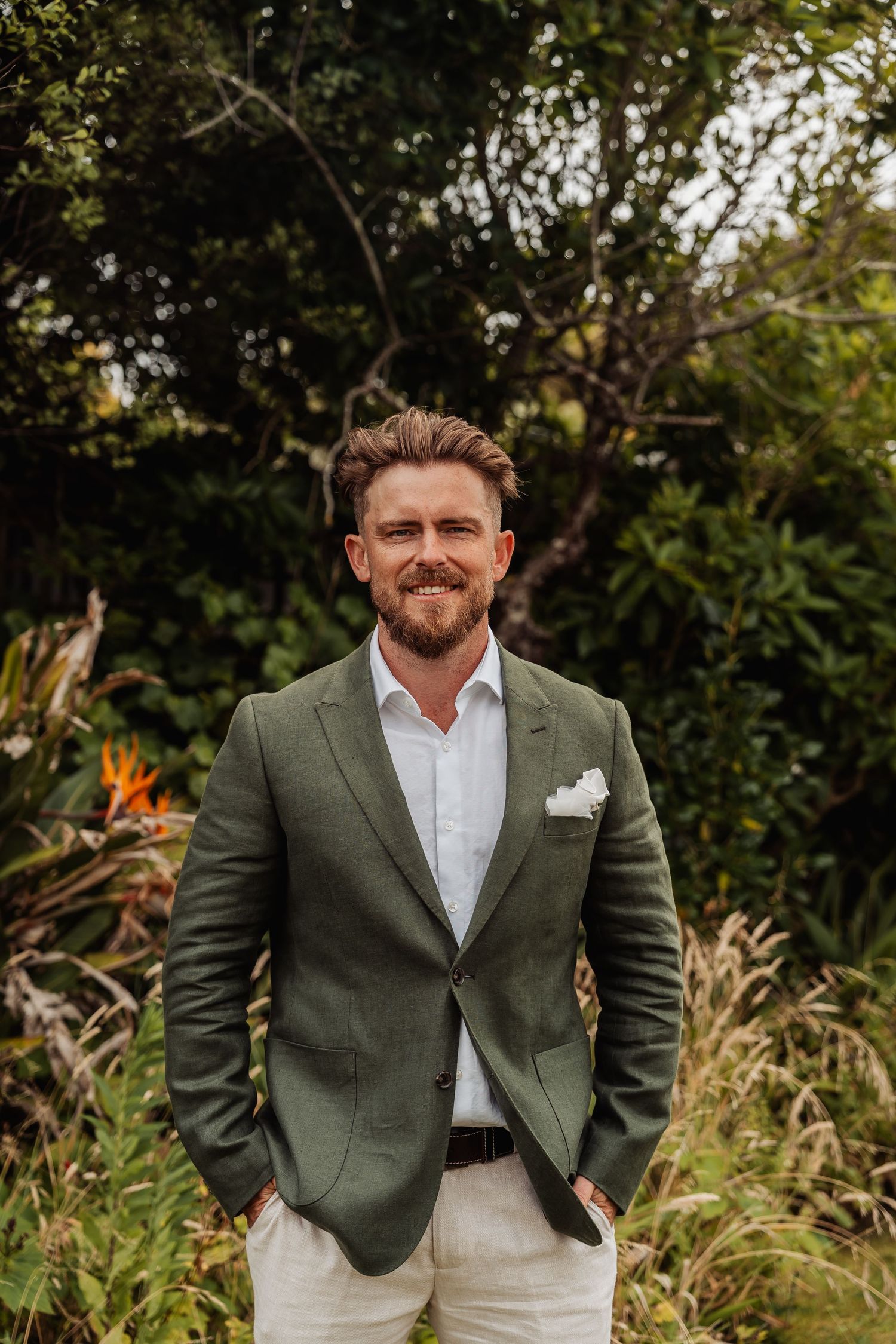 Handsome bearded groom smiling confidently in a green suit with white pocket square outdoors.
