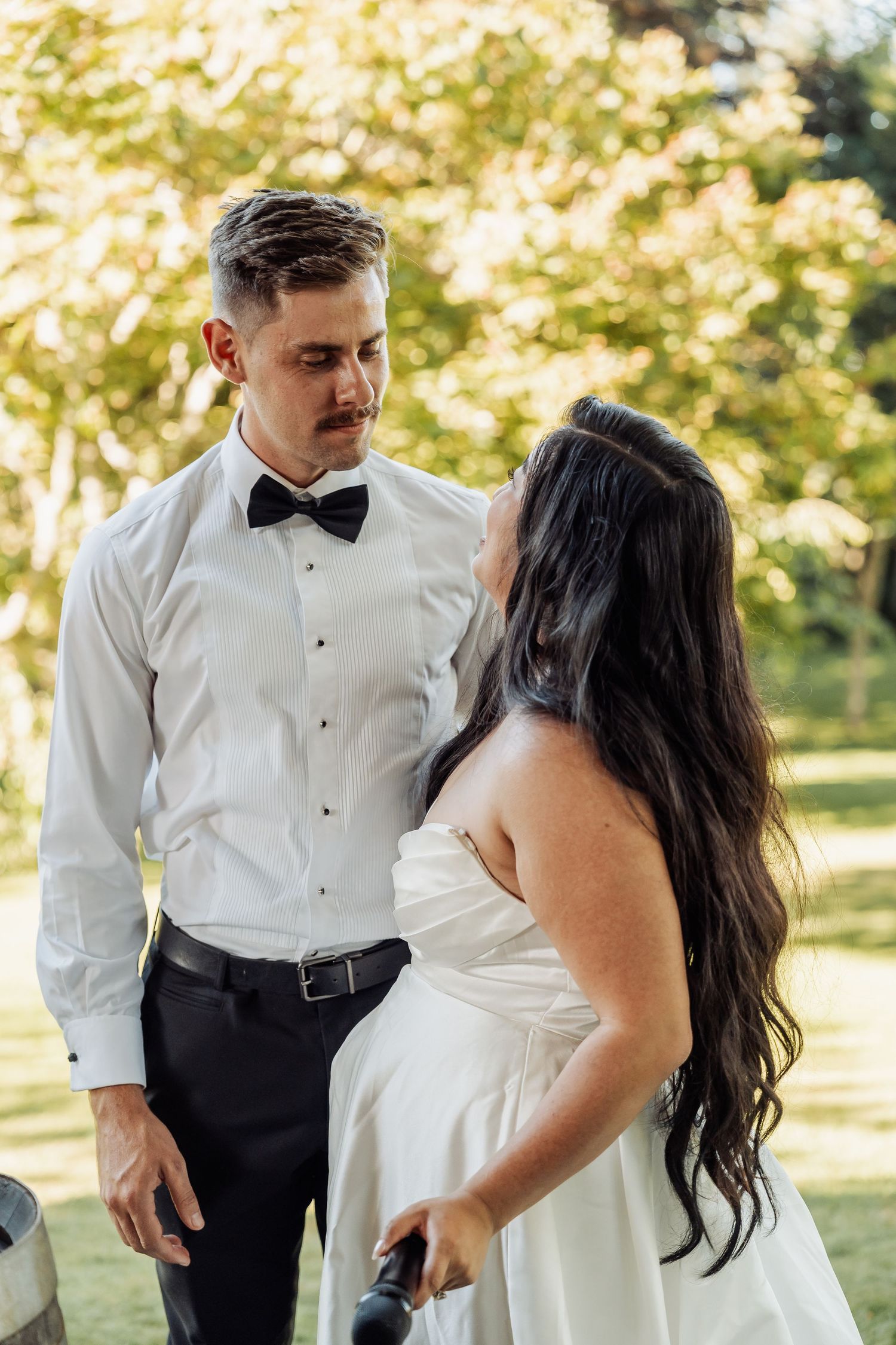 Groom in white shirt and bow tie gazes at dark-haired bride in sunny garden.