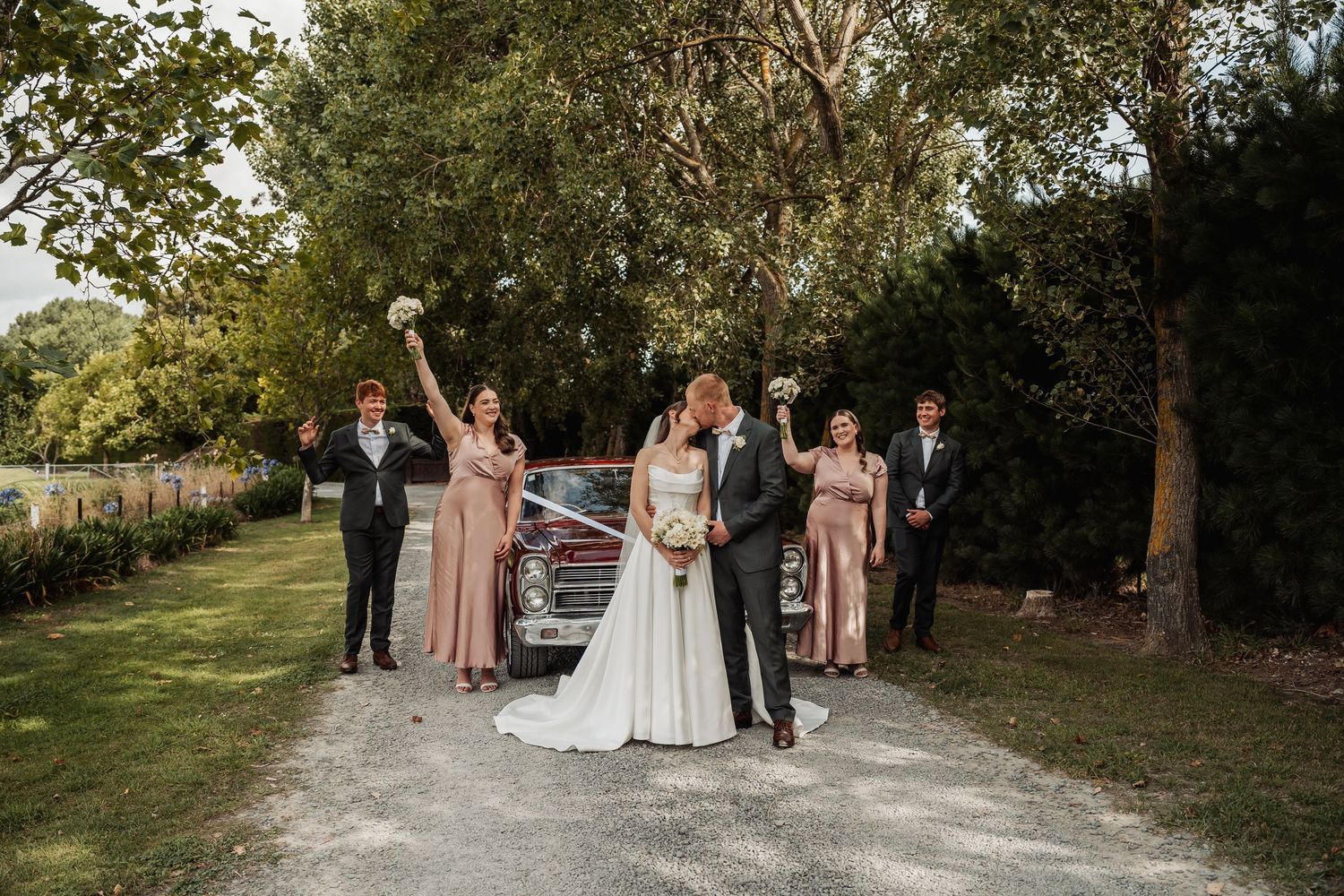Wedding party celebrates with bride and groom kissing in front of vintage car on a tree-lined gravel driveway.