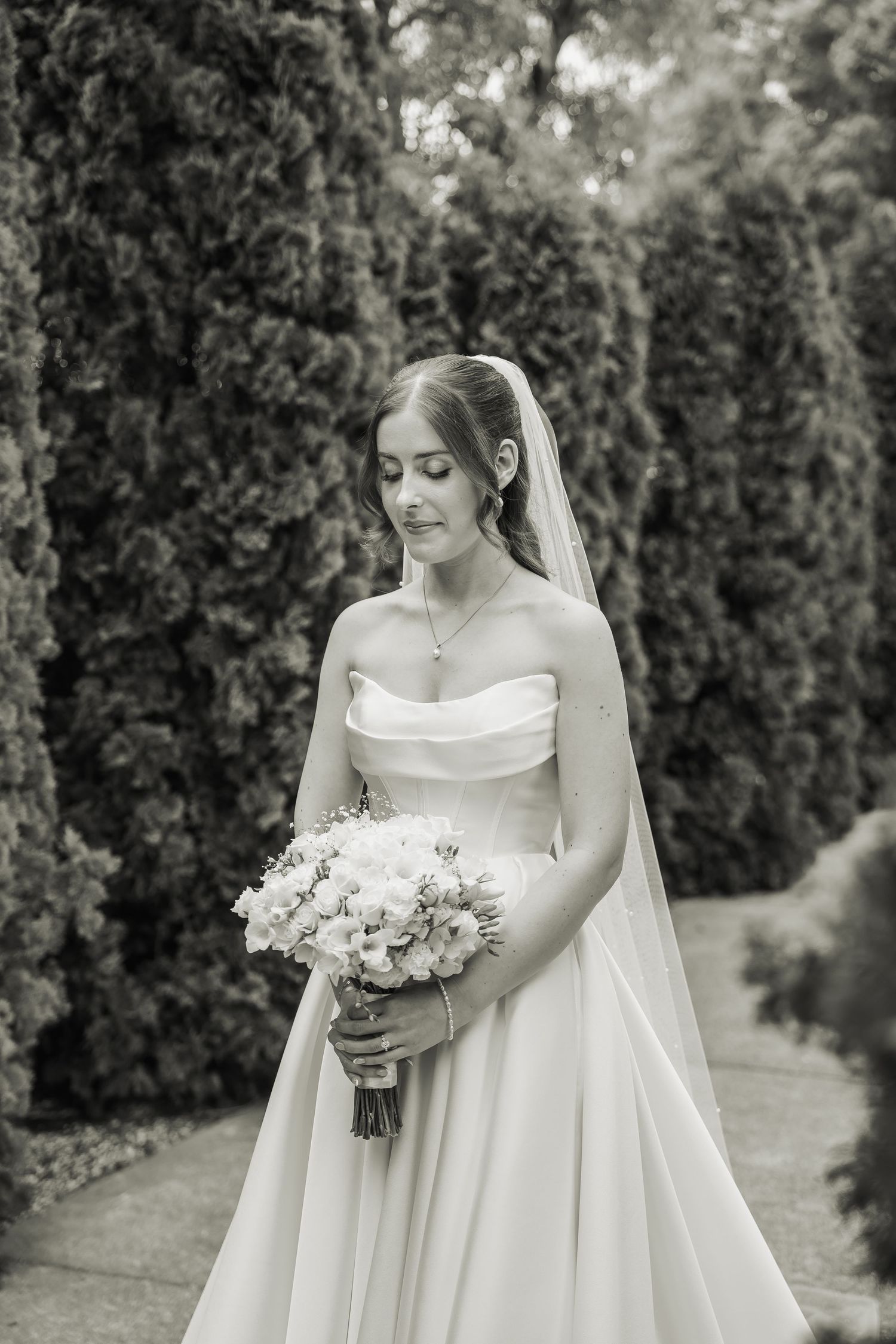 Elegant bride in strapless white gown holds bouquet among tall cypress trees in black and white photo.