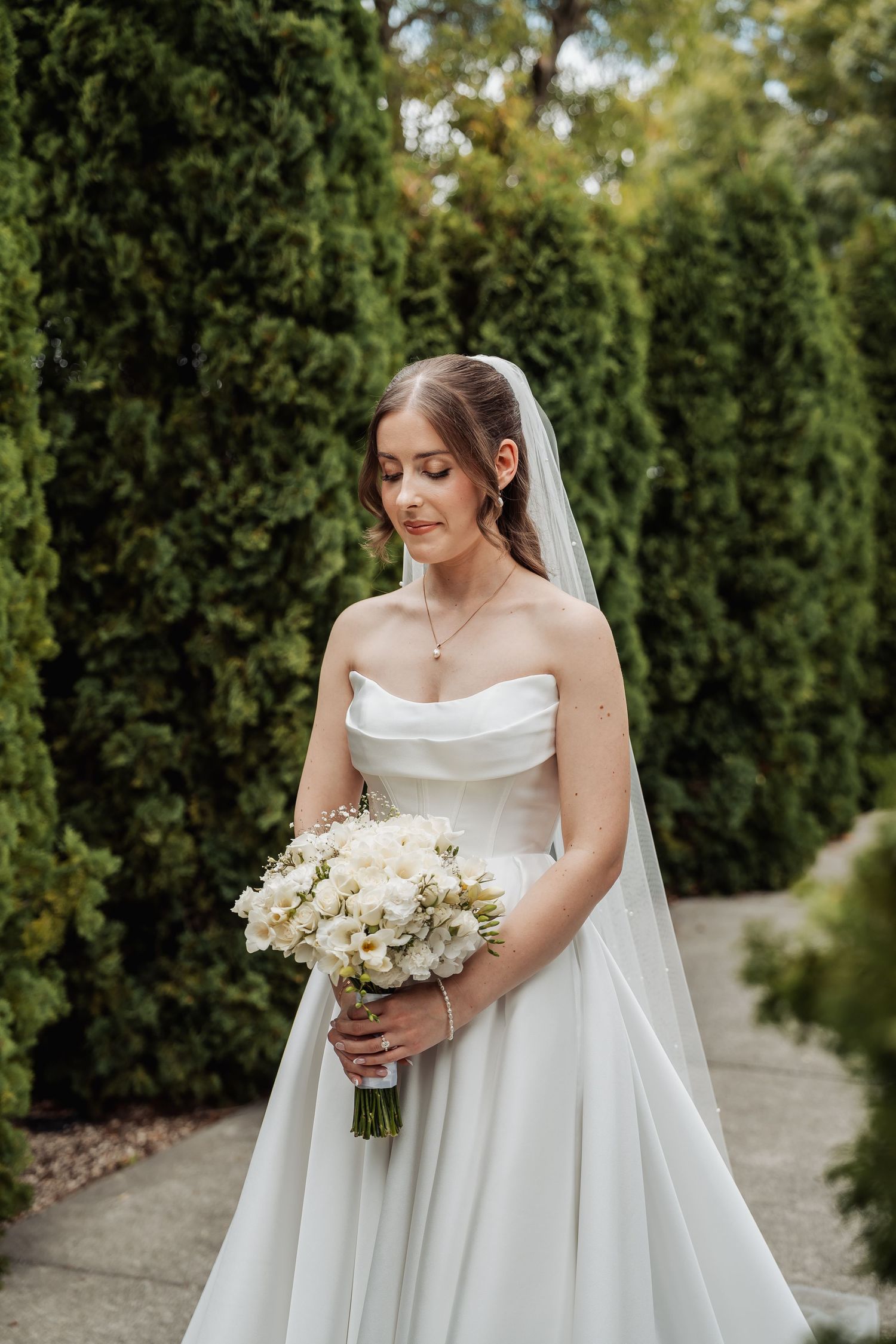 Elegant bride in strapless white gown holds a white bouquet while standing among tall green hedges.