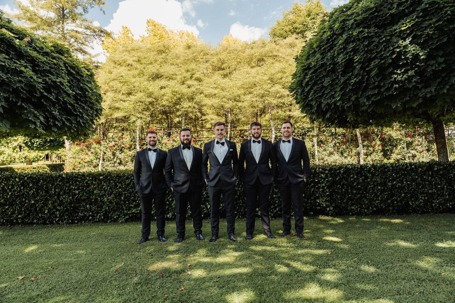 Six groomsmen in black tuxedos with bow ties posing outdoors on lush green lawn with manicured hedges and trees.