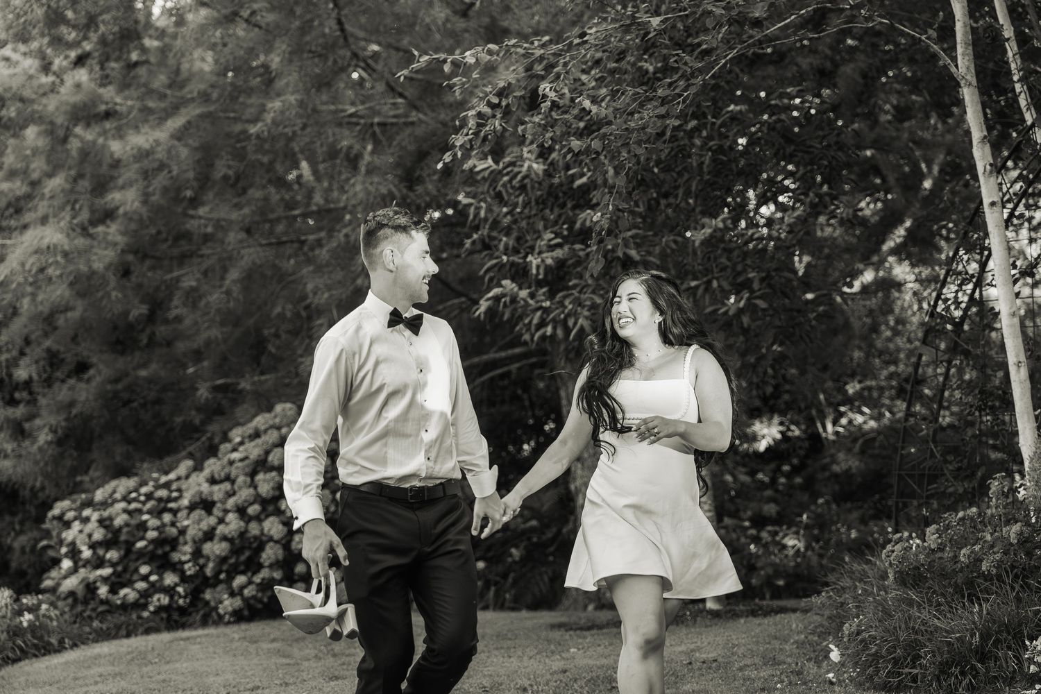 Black and white photo of bride and groom holding hands and smiling while walking through a garden.