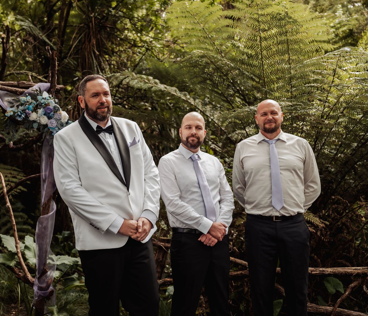 Three groomsmen in formal attire stand together at an outdoor woodland wedding ceremony.