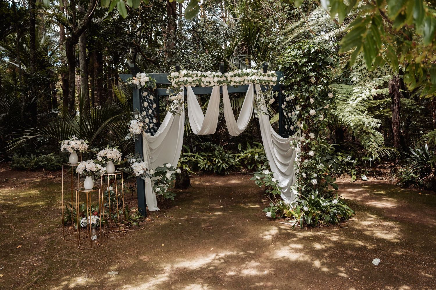 Elegant outdoor wedding arch with white draping, floral arrangements, and lush greenery in a forest setting.