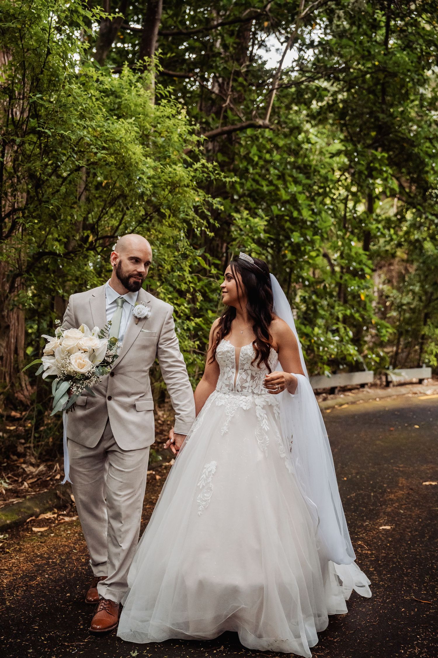 Bride and groom walk hand in hand along tree-lined path in lush garden setting.