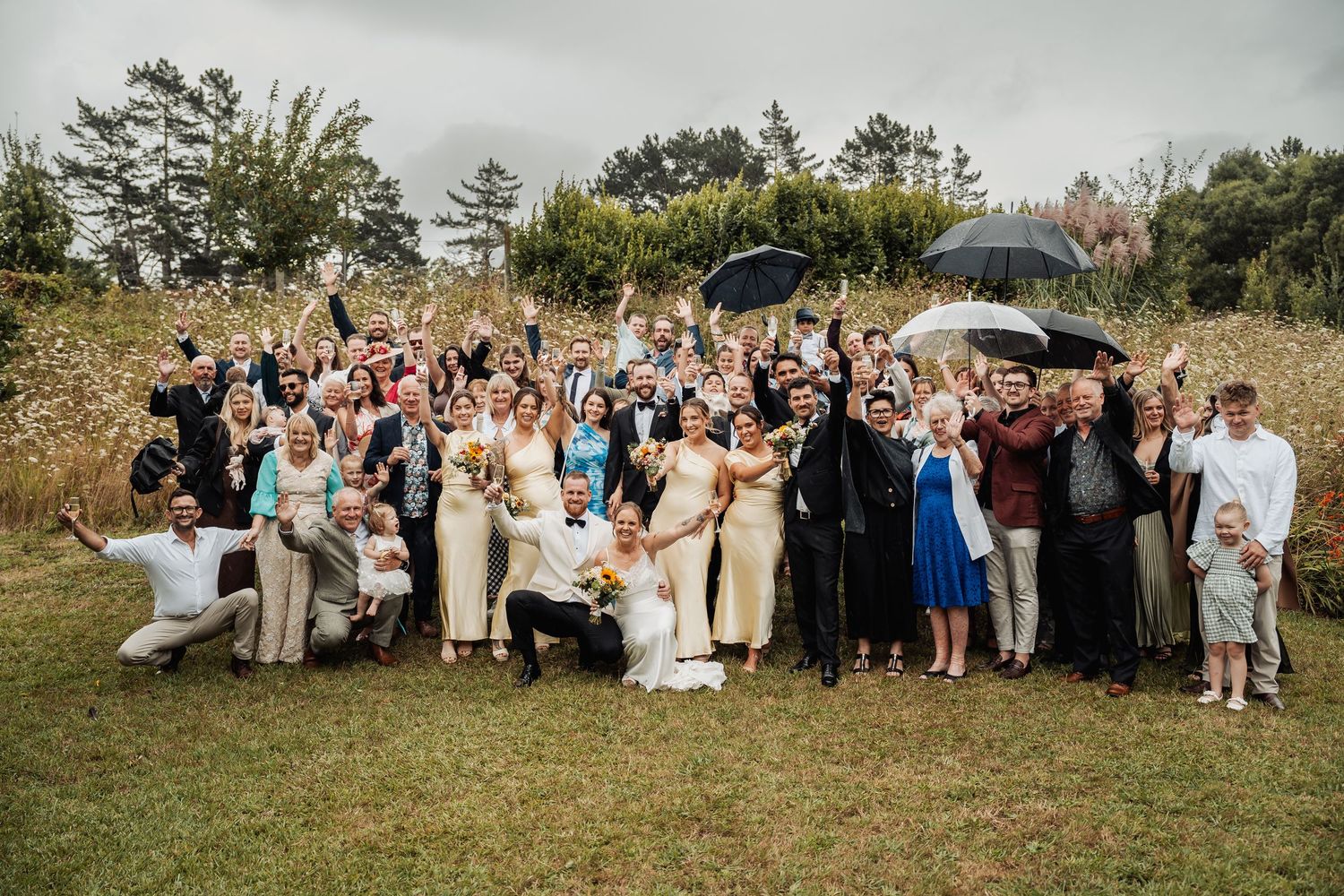 Large wedding group photo with guests cheering and holding umbrellas on overcast day.