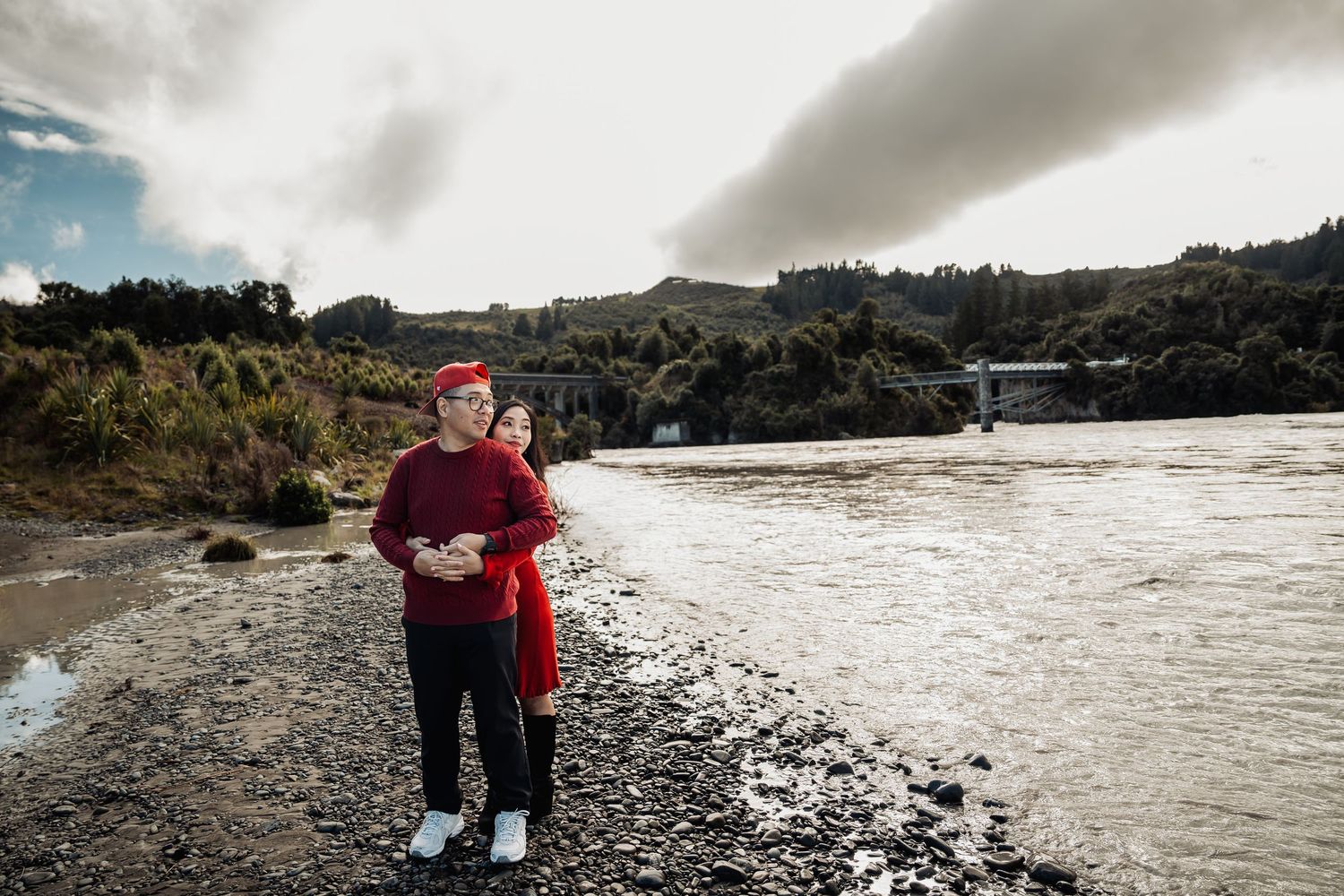 Couple in red outfits embracing while standing on a rocky riverbank with stormy skies and a bridge behind them.