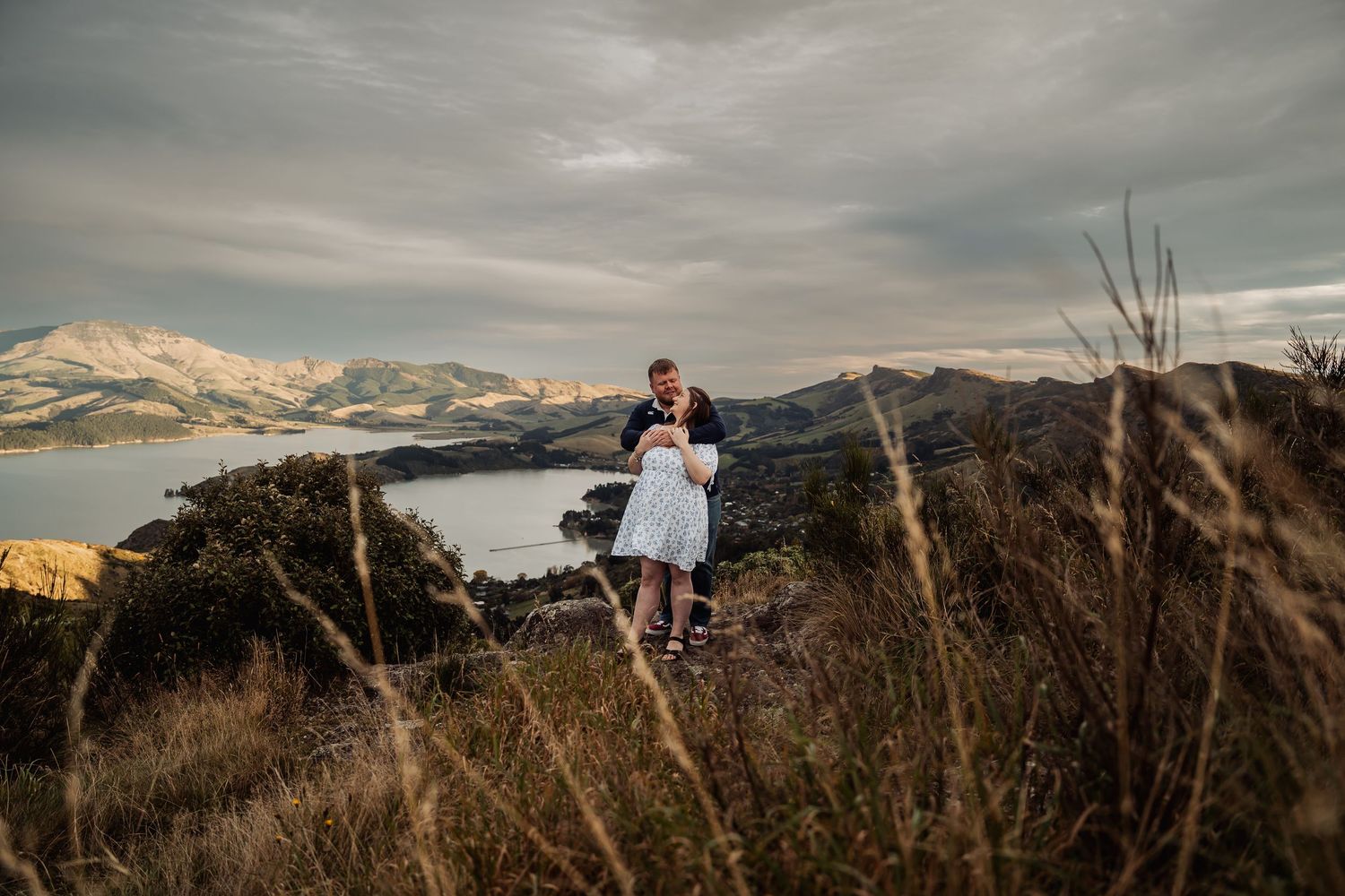 Couple embracing on a hilltop with stunning lake and snow-capped mountain views under a dramatic sky.