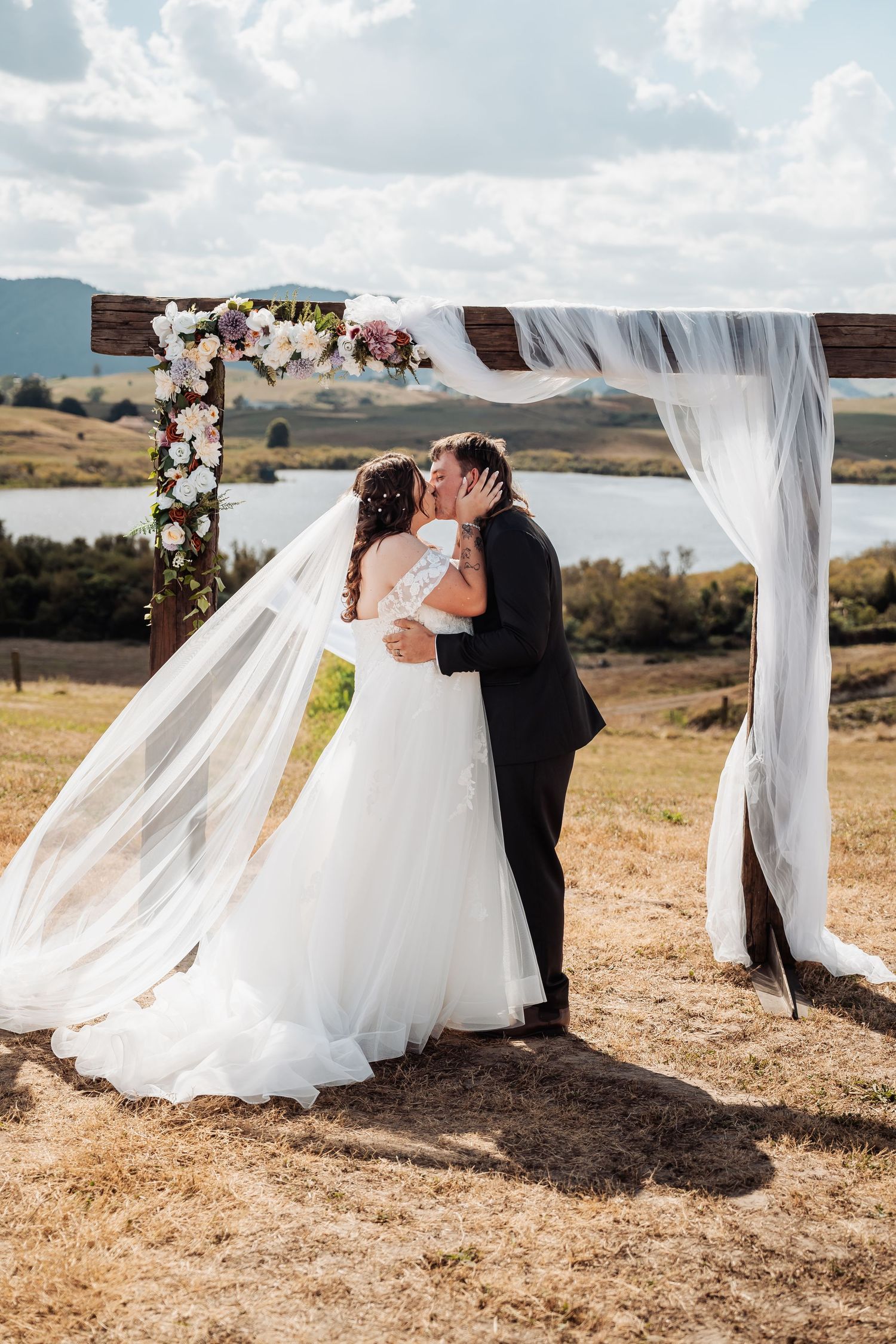 Bride and groom share first kiss under floral wooden arch with flowing veil and lake views behind them.