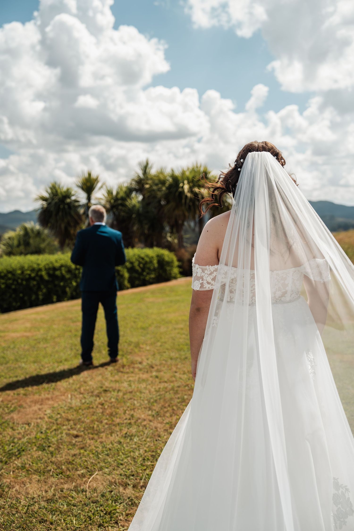 Bride in white gown and veil walks toward groom on green lawn with palm trees and mountains behind.