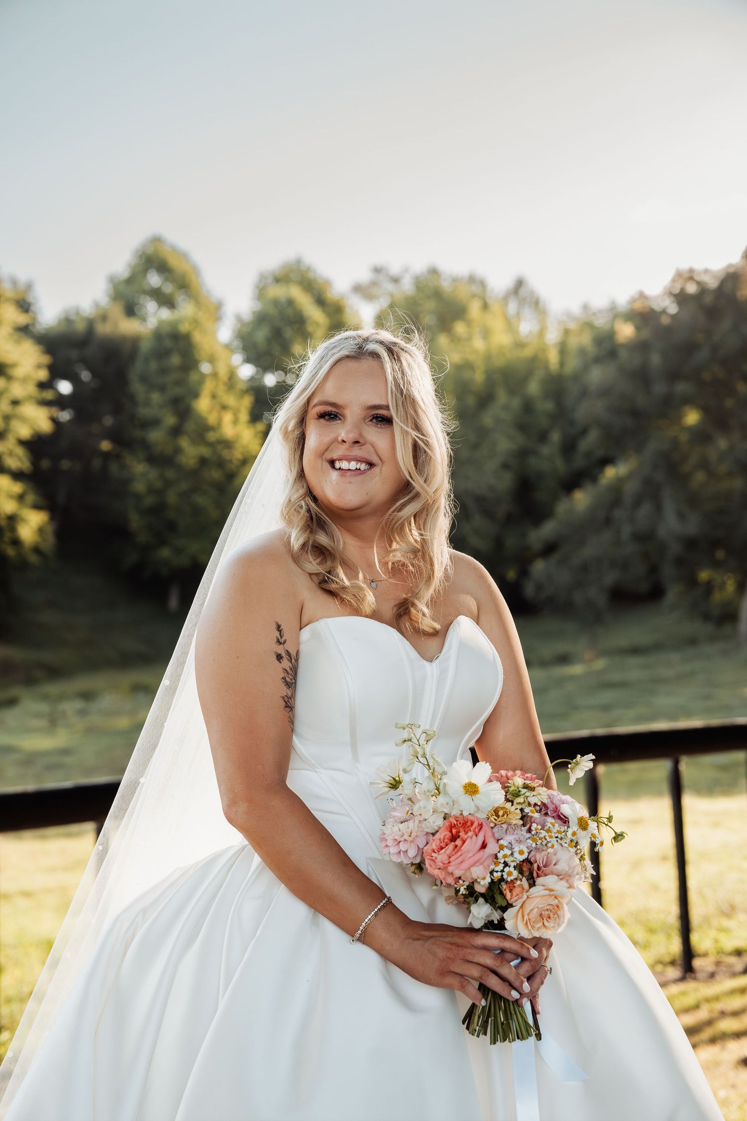 Smiling bride in strapless white gown holds a colorful bouquet of pink and white flowers outdoors.