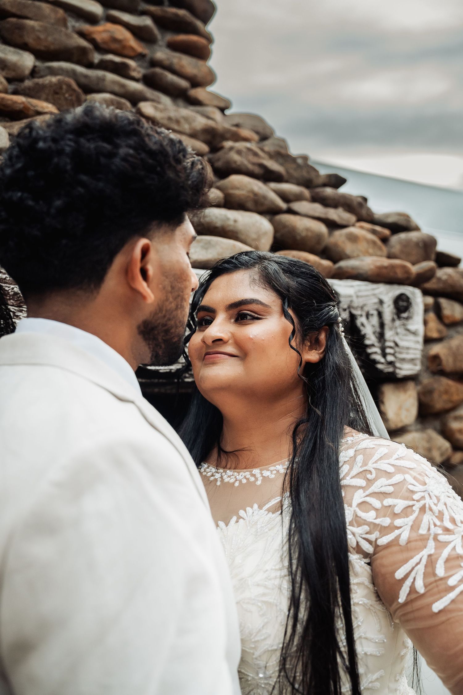 Bride in lace wedding gown gazes lovingly at groom against rustic stone wall backdrop outdoors.