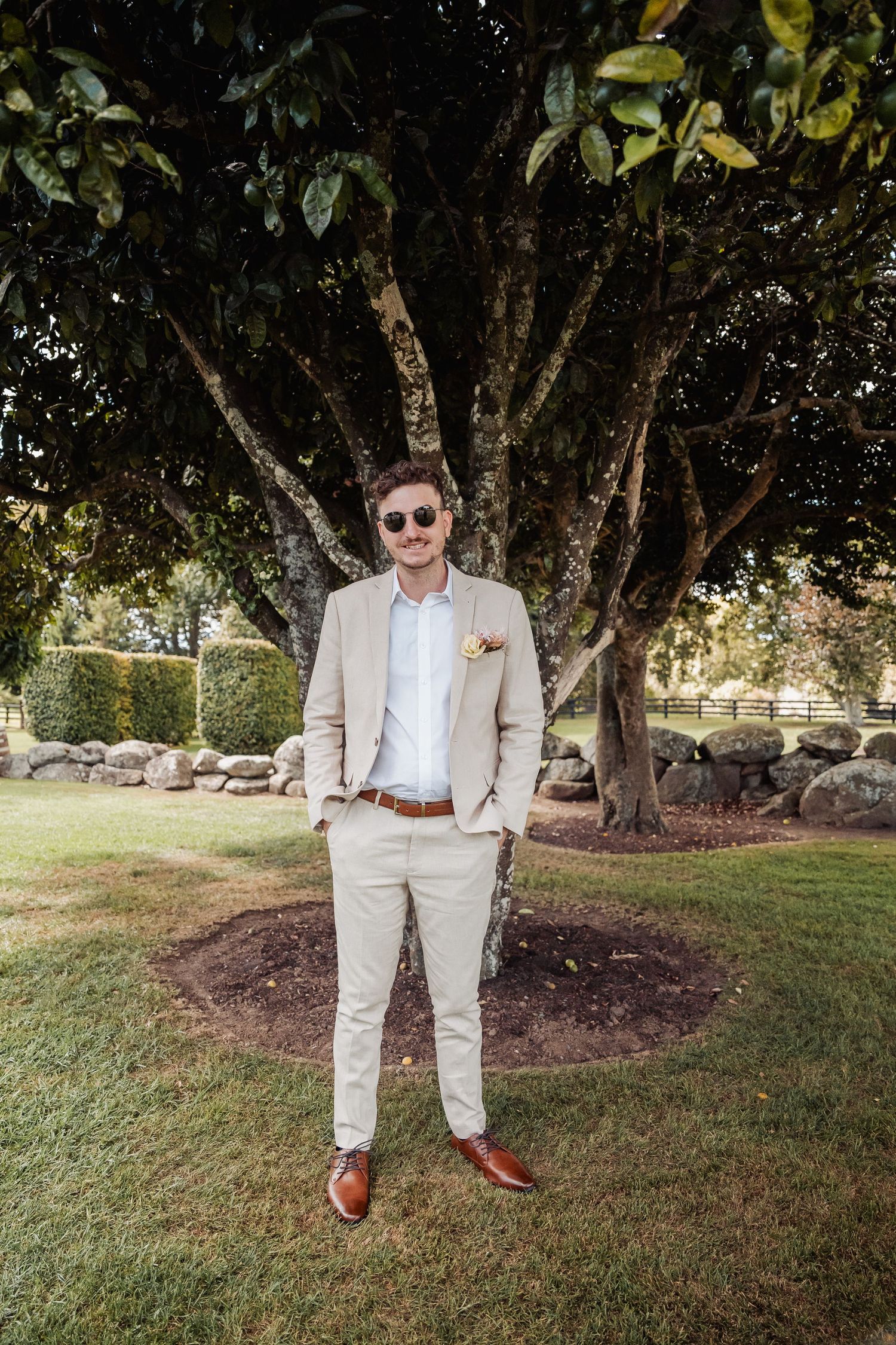 Groom in beige suit and sunglasses standing confidently under a large tree.