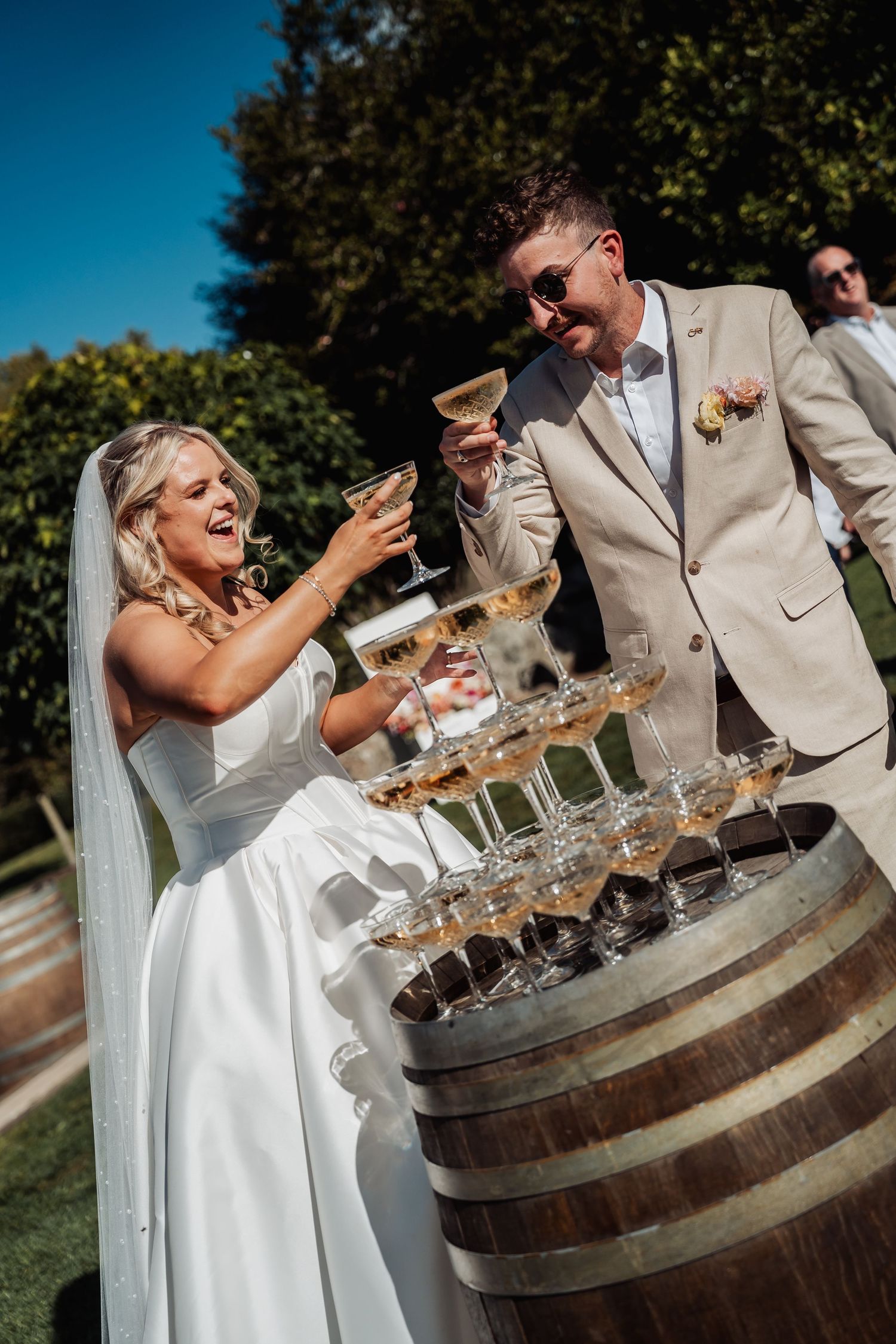 Bride and groom toasting champagne glasses over a barrel tower of glasses at outdoor wedding.