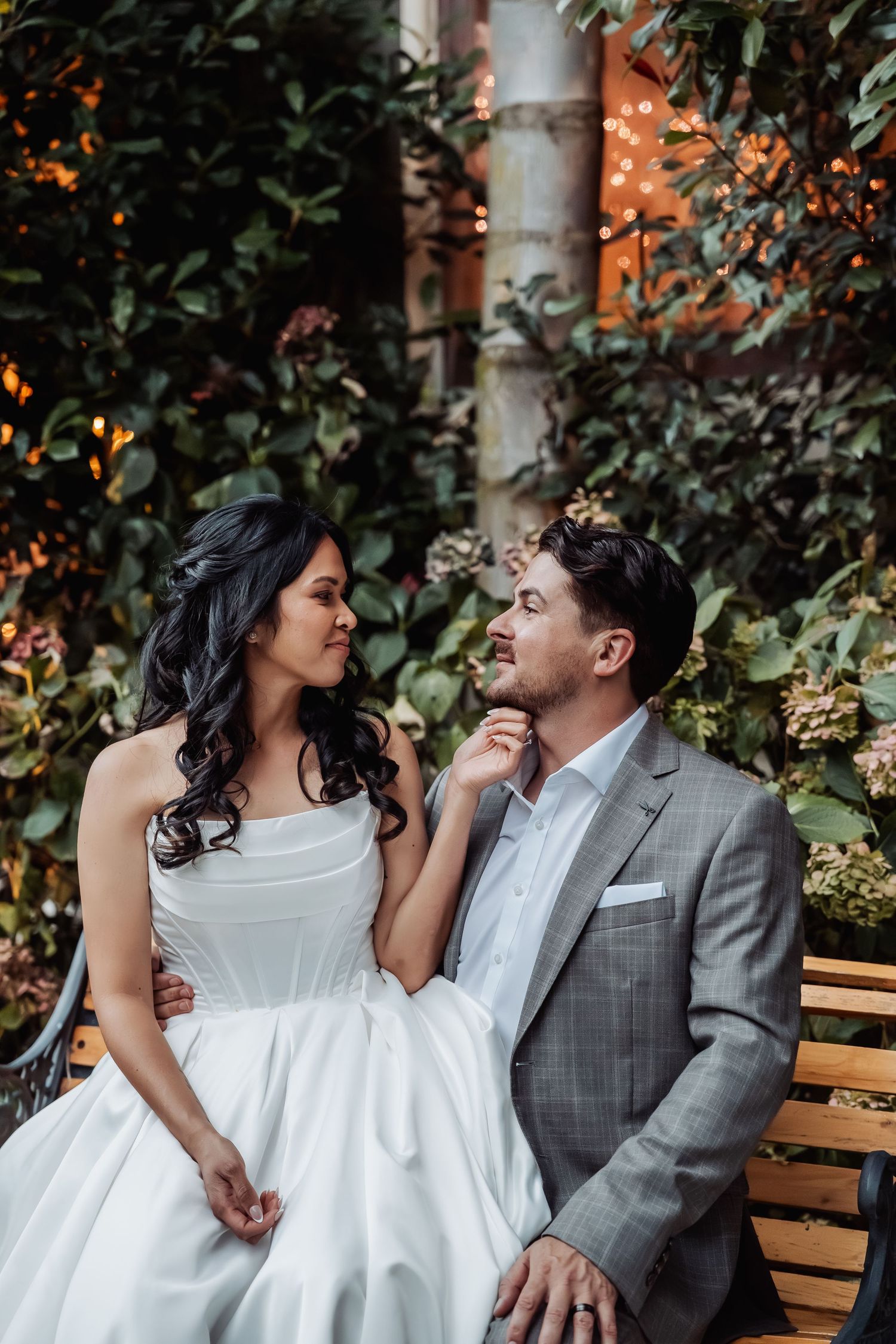 Bride and groom sit on bench gazing at each other surrounded by lush greenery and soft bokeh lights.