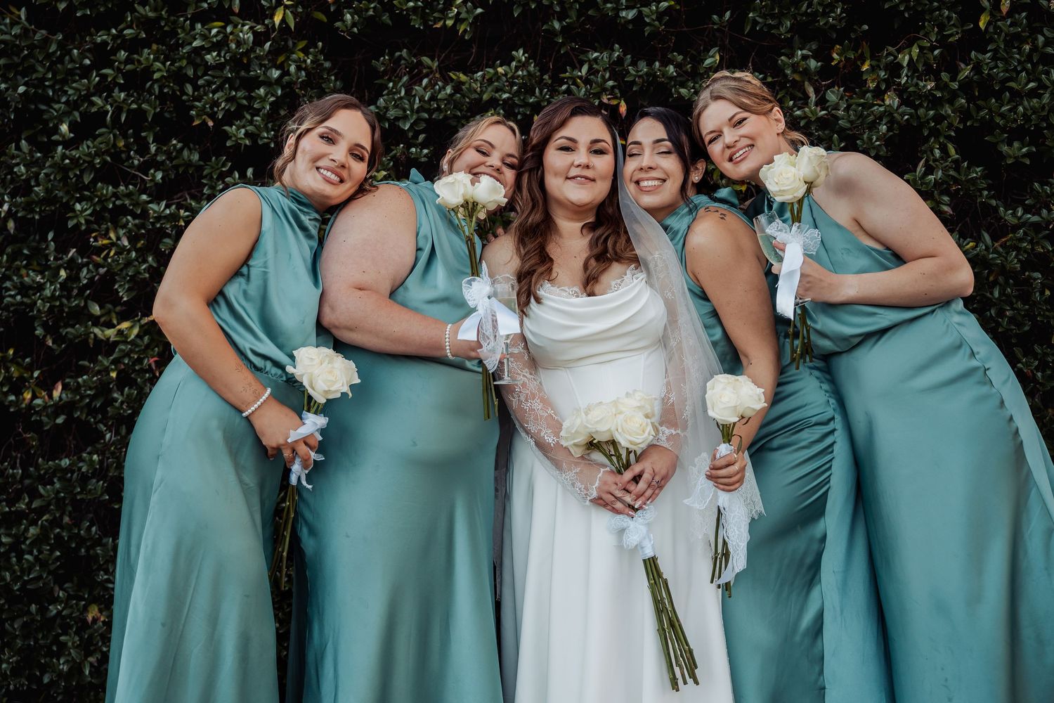 A bridal party wearing matching teal bridesmaid dresses poses together with the bride in an outdoor garden setting.