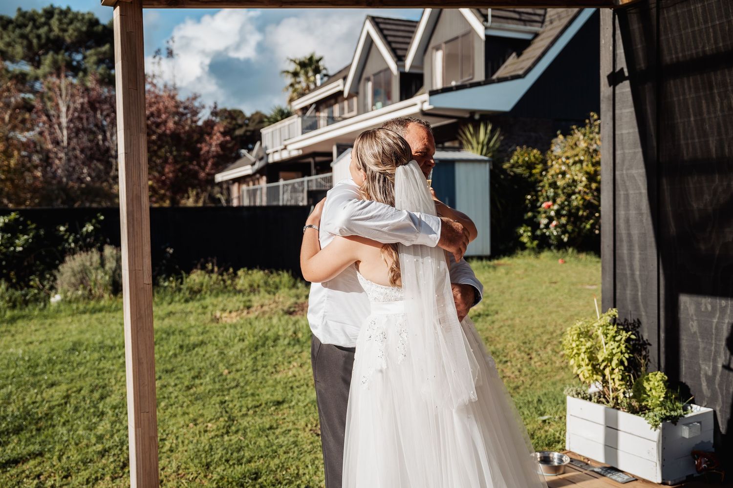 Newlyweds share an intimate embrace on their wedding day in front of a rustic barn with palm trees and greenery.
