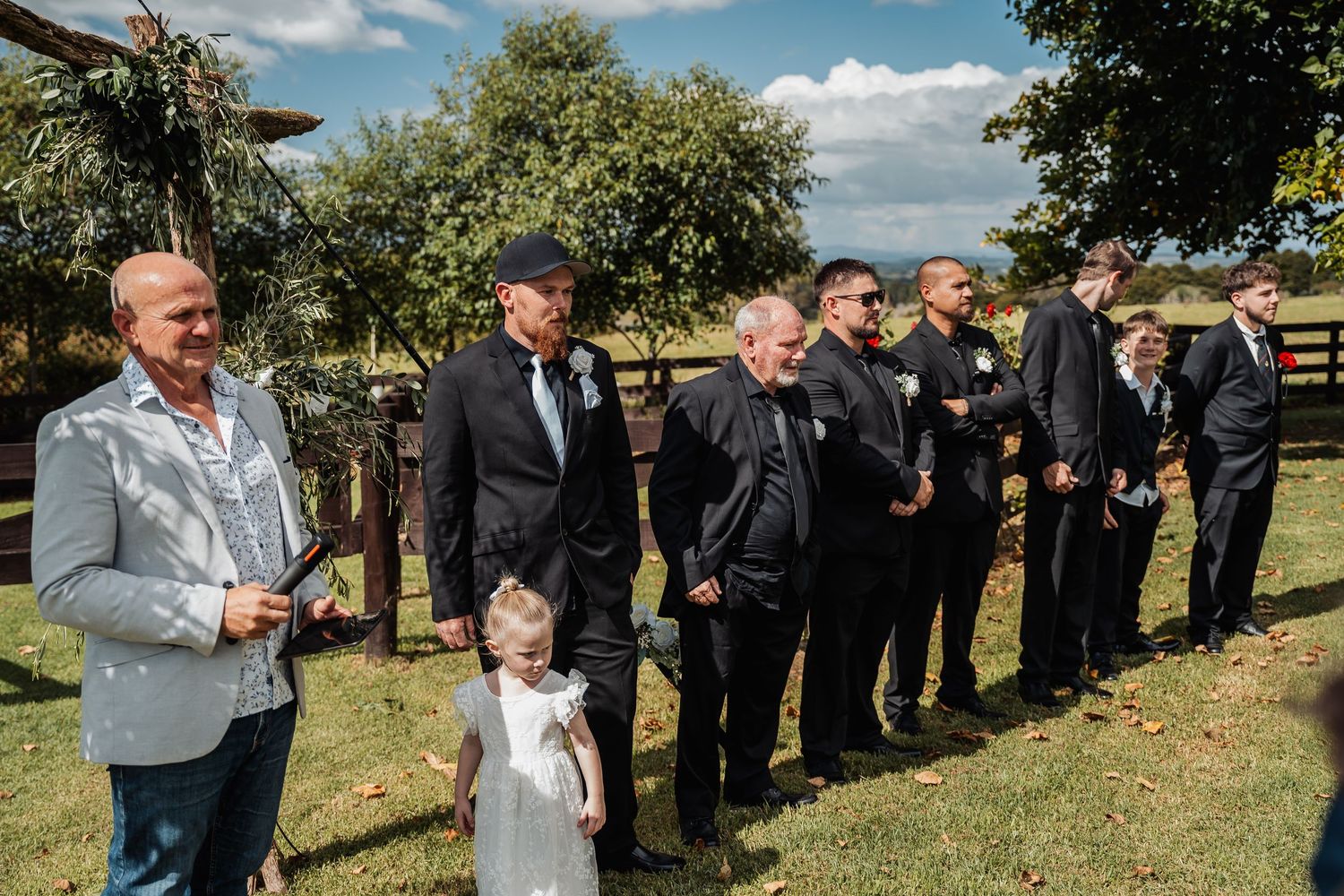 A wedding party stands in a line on a sunny lawn with groomsmen in dark suits and a little flower girl.