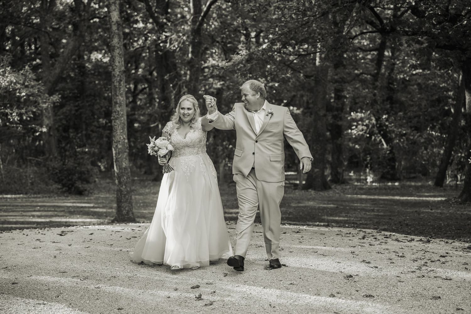 Bride and groom walking joyfully hand in hand on gravel path in black and white wedding photo.