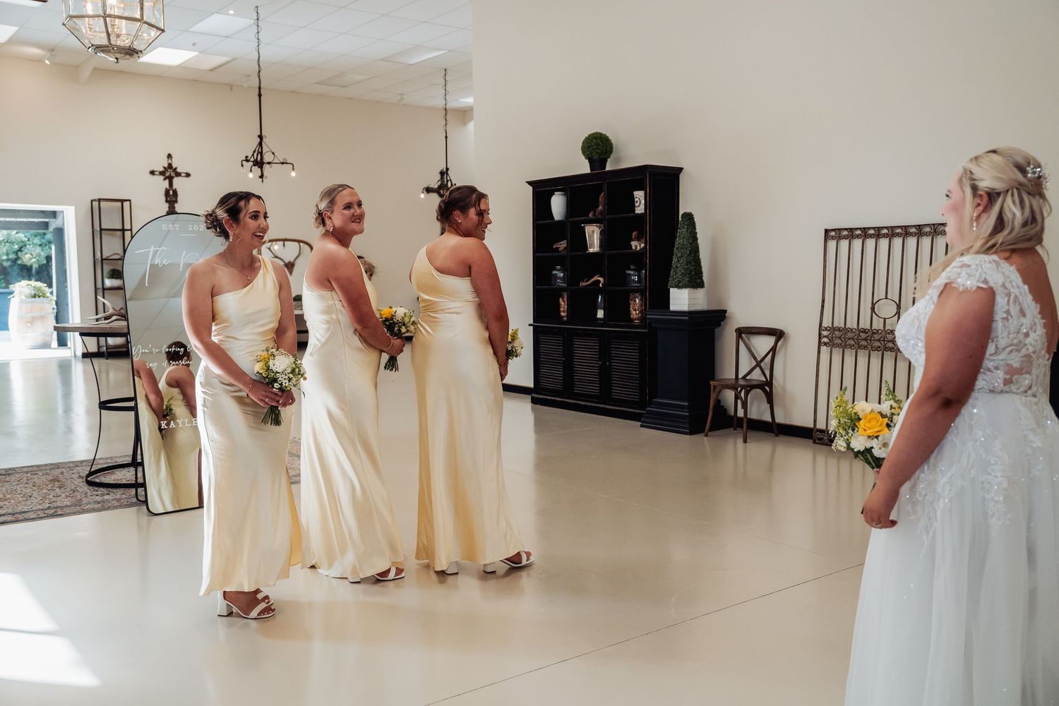 Bride in white gown shares a joyful first look moment with three bridesmaids in ivory dresses holding bouquets.