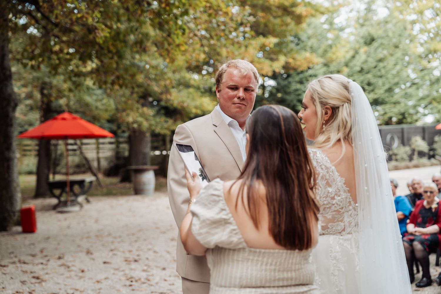 Bride and groom exchanging vows with officiant outdoors under trees.