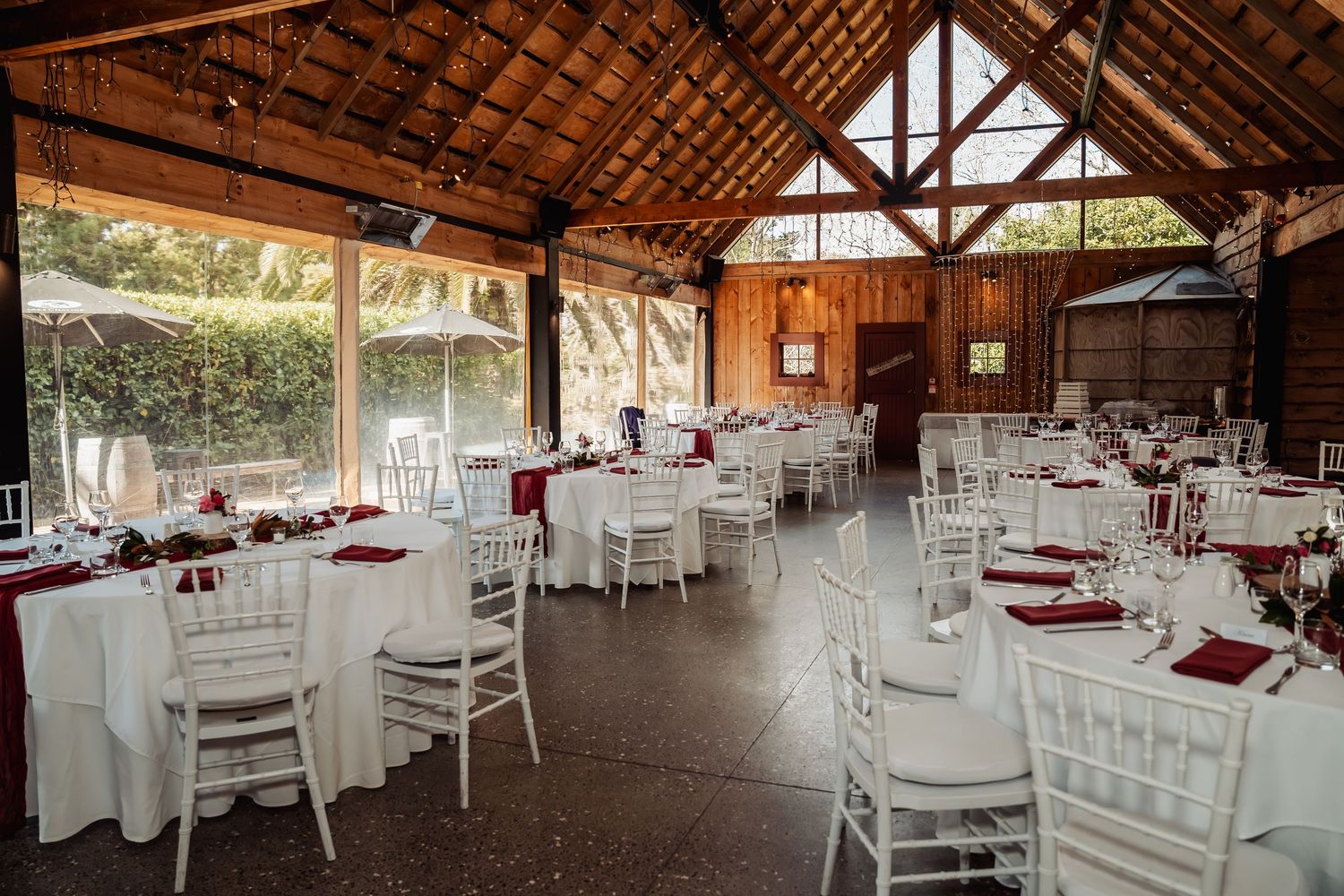 Elegant barn wedding reception venue with white Chiavari chairs, round tables with burgundy linens, and rustic wooden ceiling.