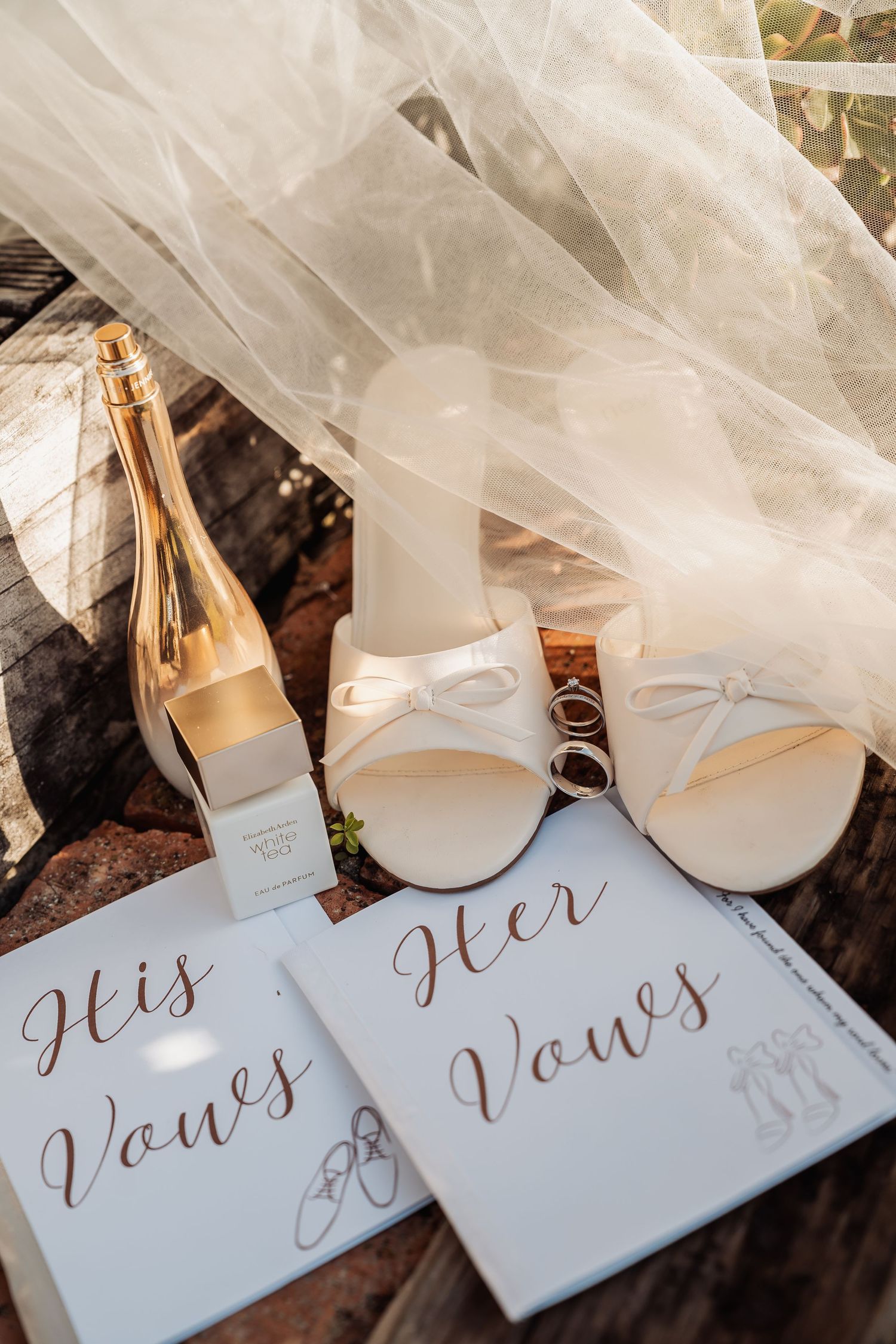 Wedding flat lay with vow books, white heels, rings, and champagne bottle under bridal veil.