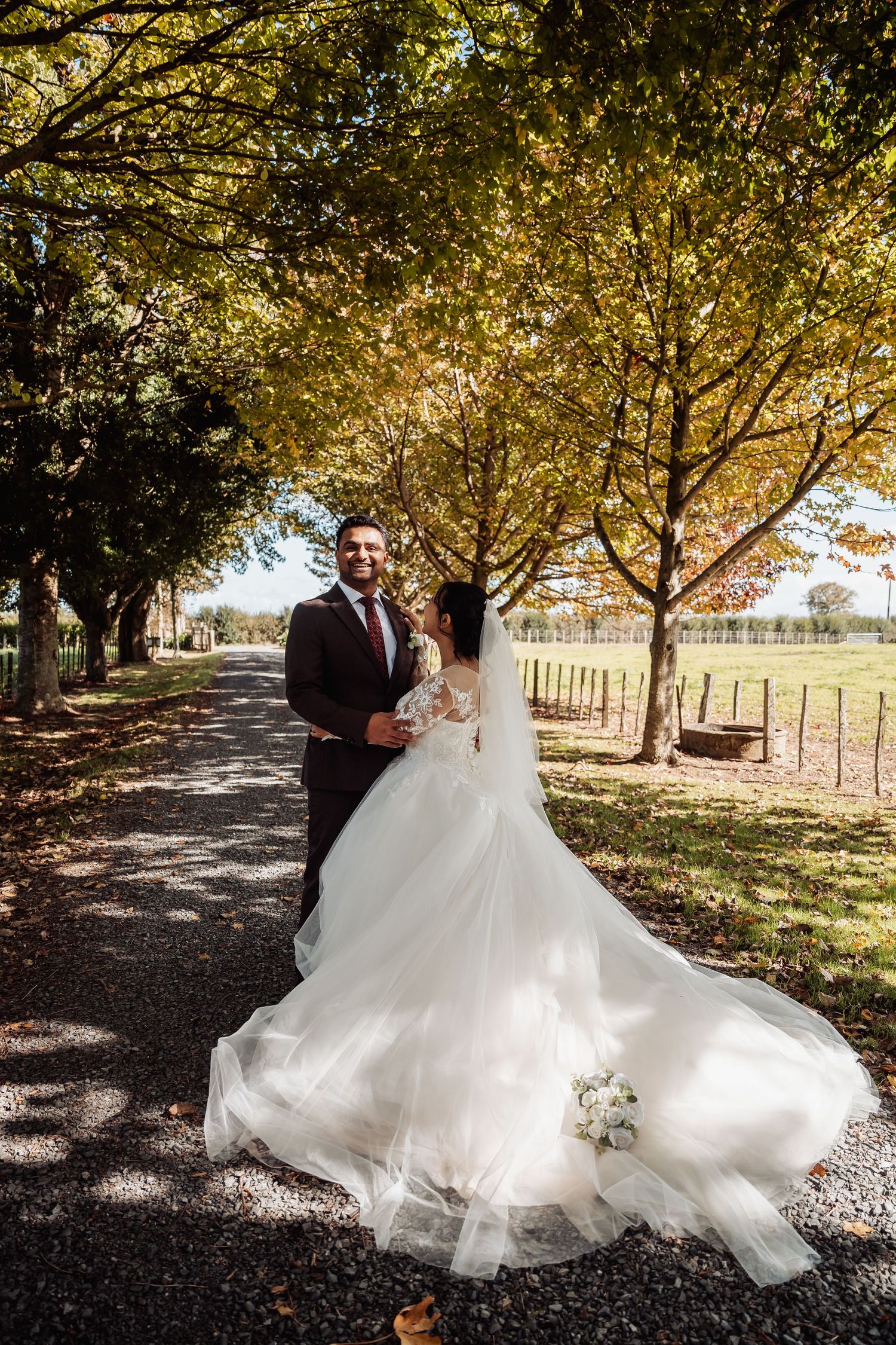 Bride and groom embrace on autumn tree-lined path, brides flowing white tulle gown trailing behind.