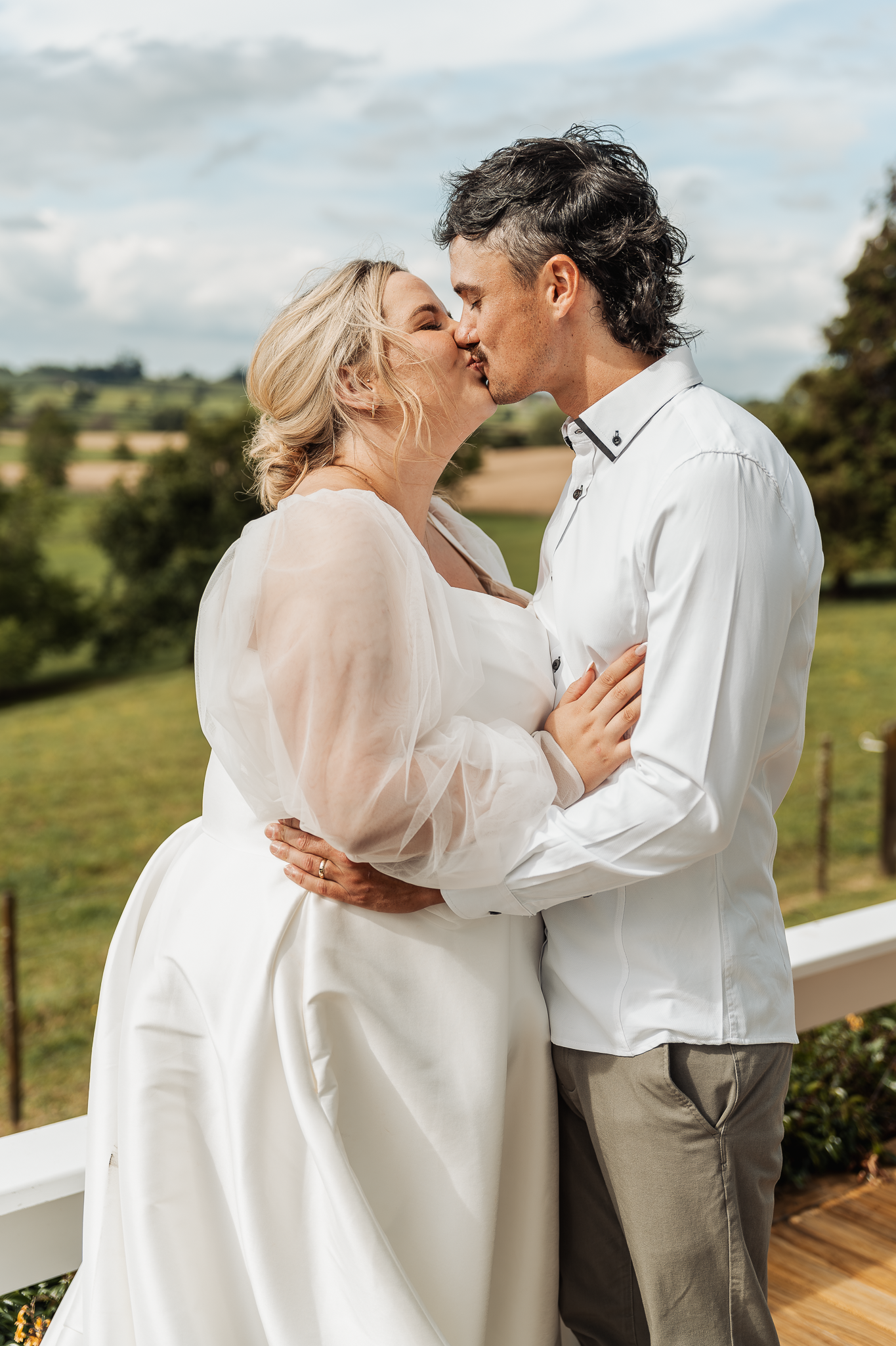 Romantic wedding couple sharing a kiss on wooden deck with scenic countryside view.