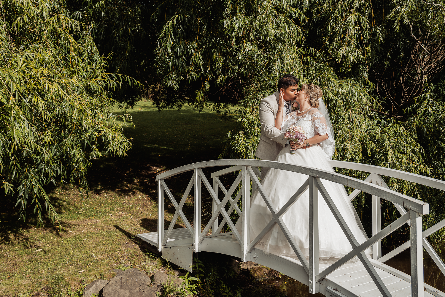 Wedding couple poses together on a decorative white bridge in a garden setting.
