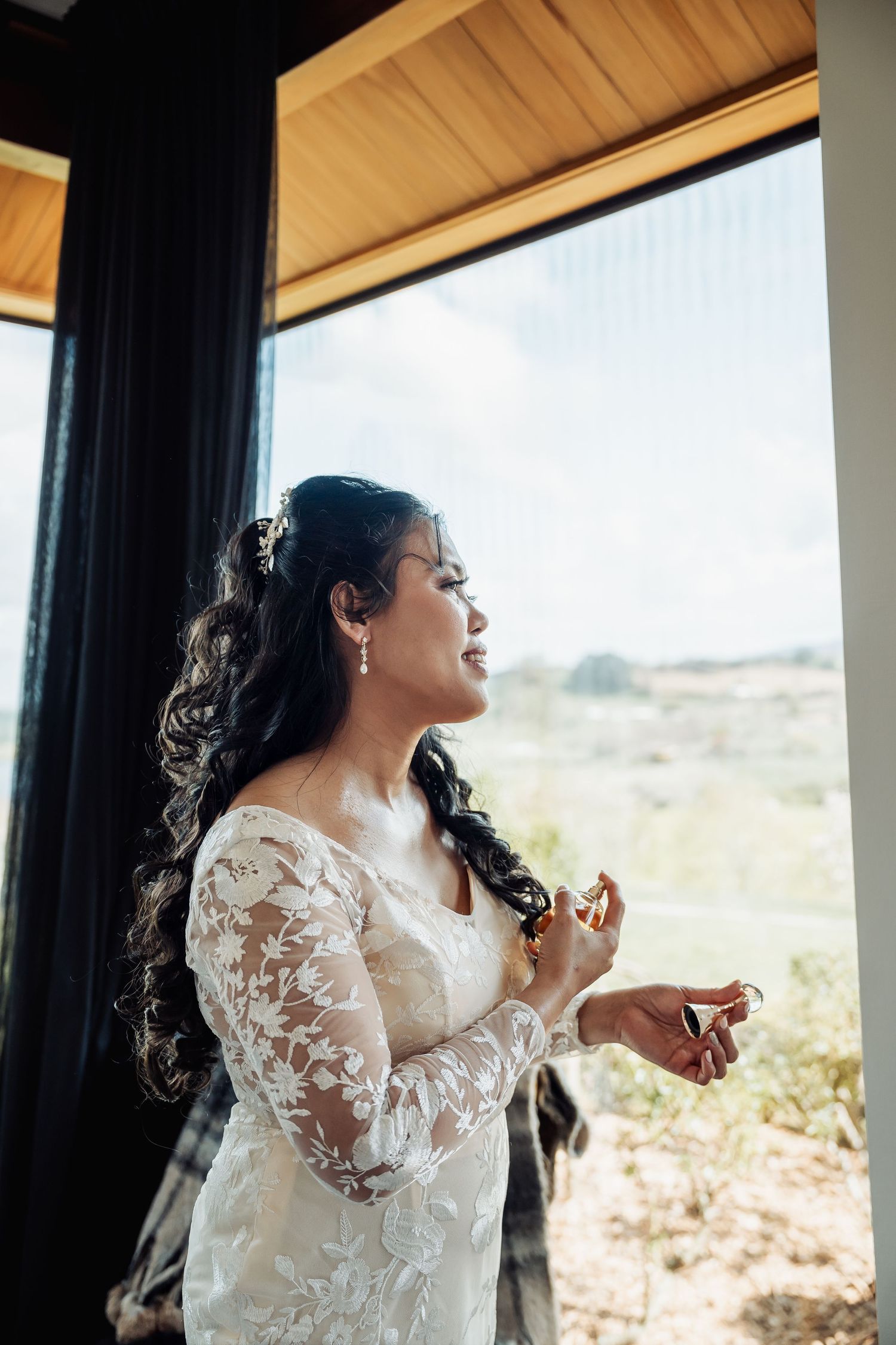 A bride in a lace wedding dress gazes out a window during preparation moments.