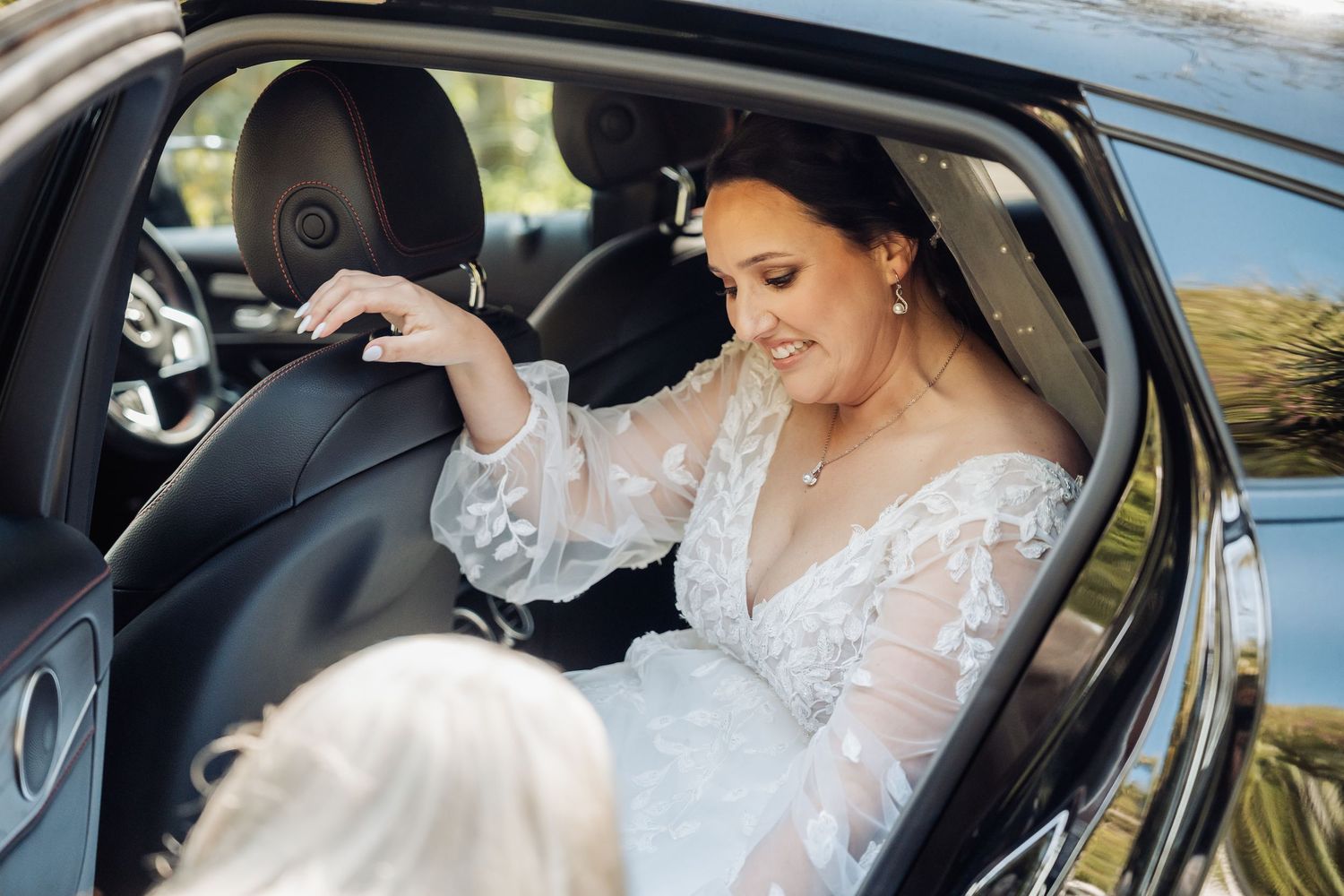 A bride in a white lace wedding dress enters a black luxury car on her special day.