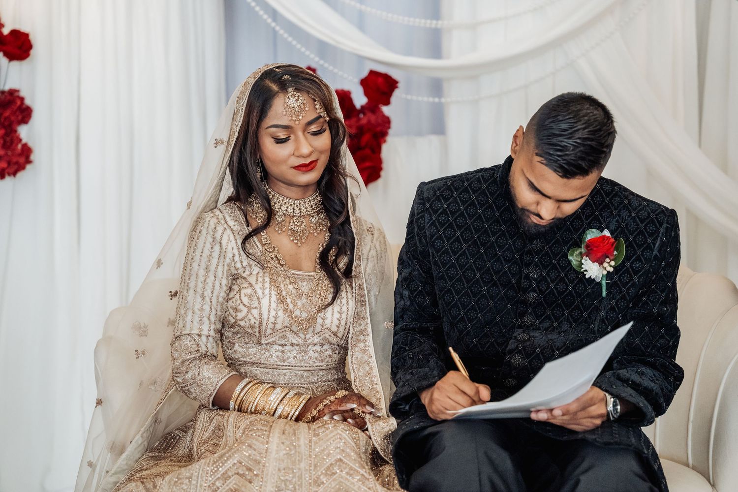 A couple signing documents together at an elegant indoor ceremony setting with red floral decor.