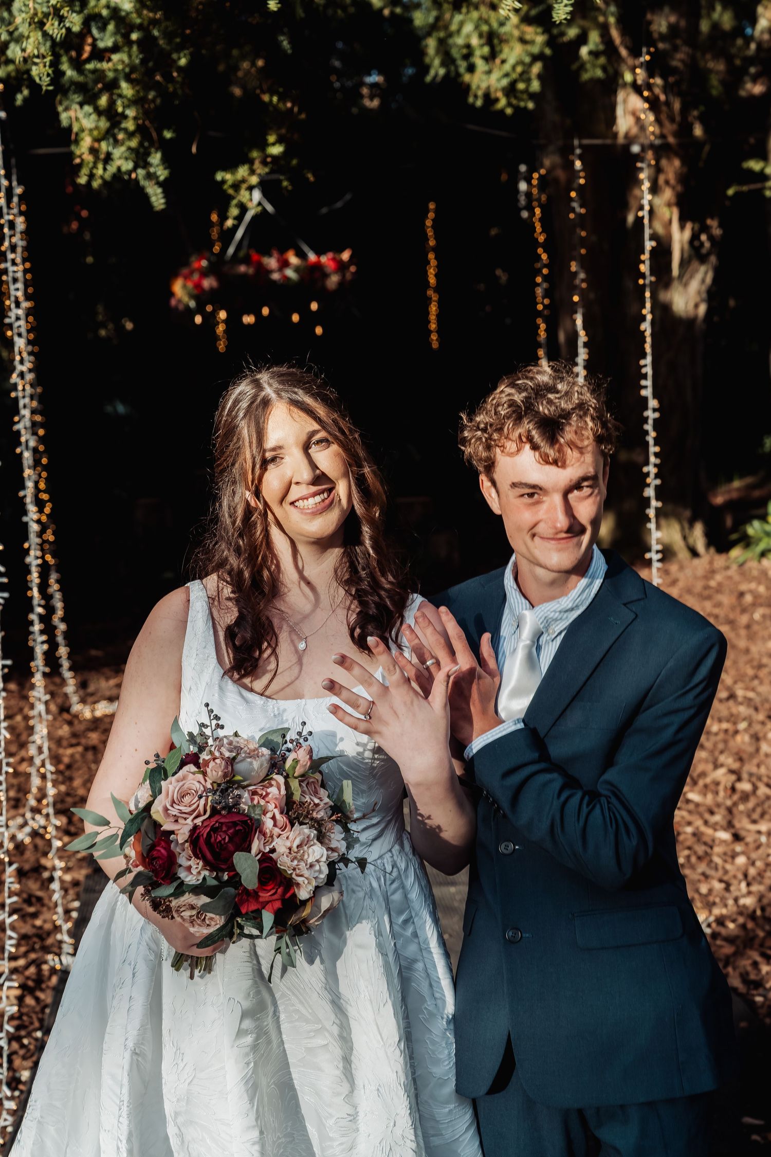 Newlyweds show off wedding rings smiling before fairy light backdrop with bridal bouquet.
