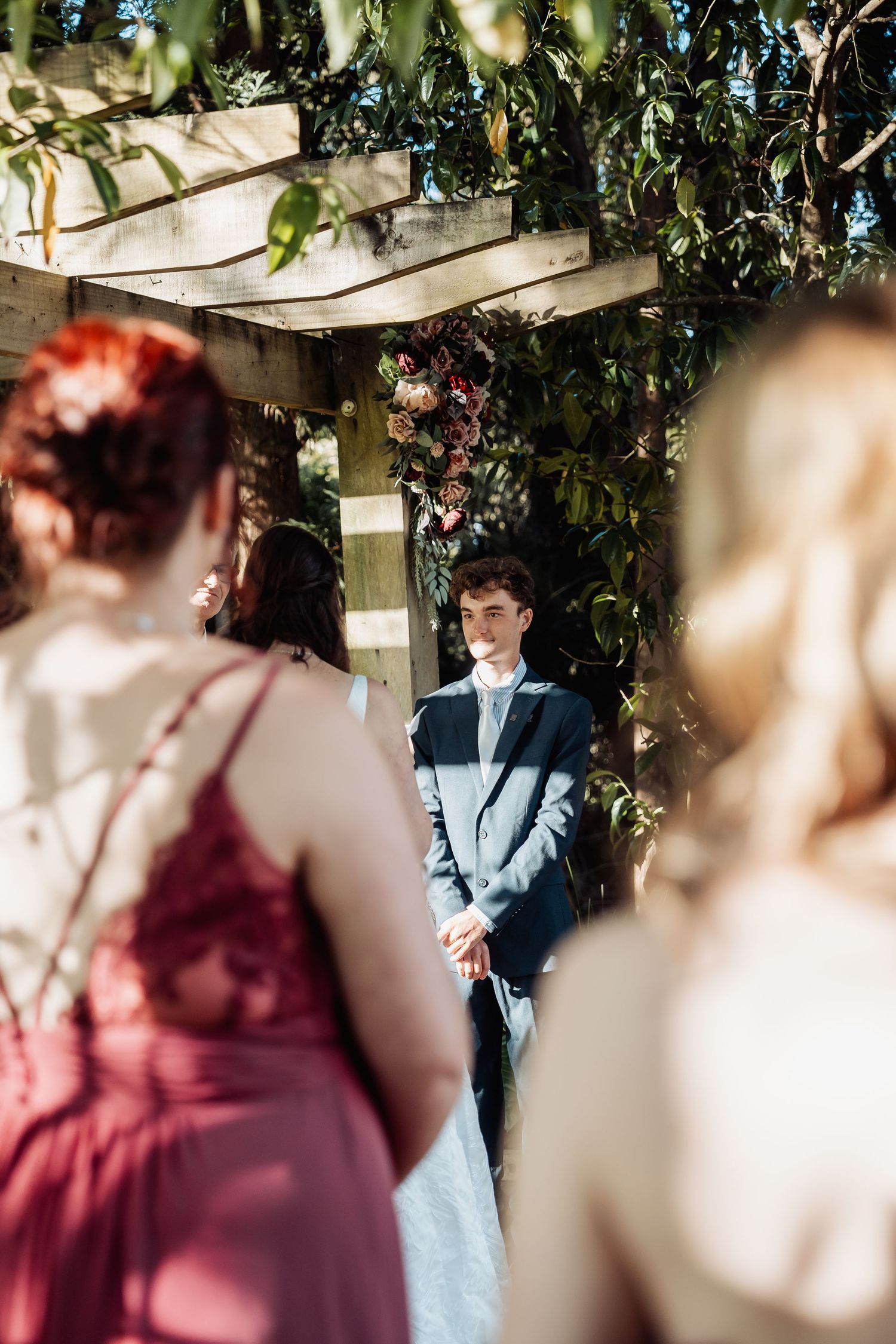 Groom stands at floral altar watching bride approach during outdoor wedding ceremony.
