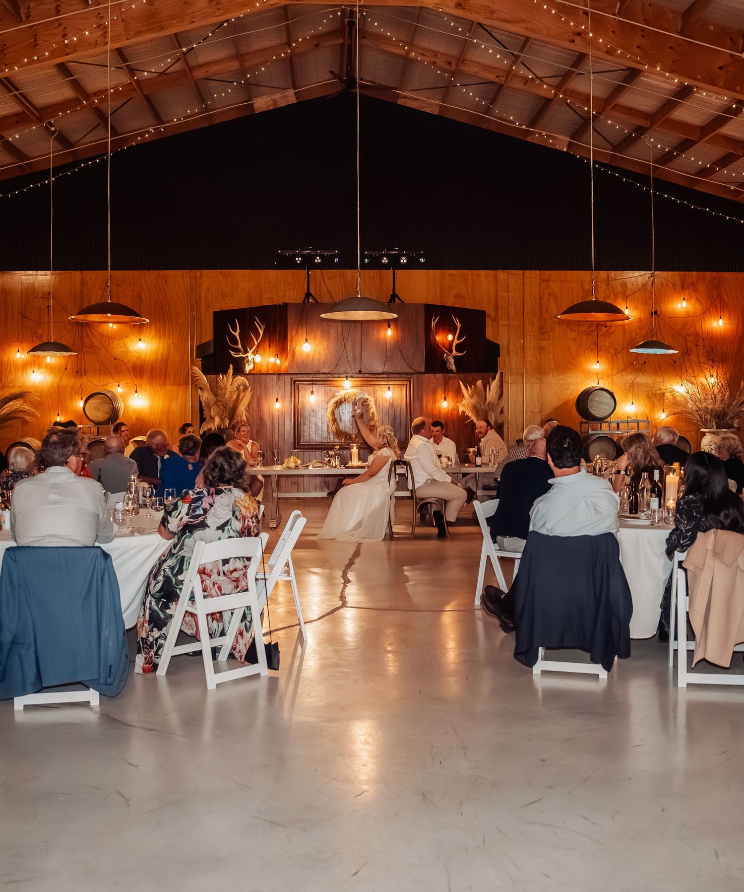 Elegant barn wedding reception with string lights, pendant lamps, and guests seated at round tables.